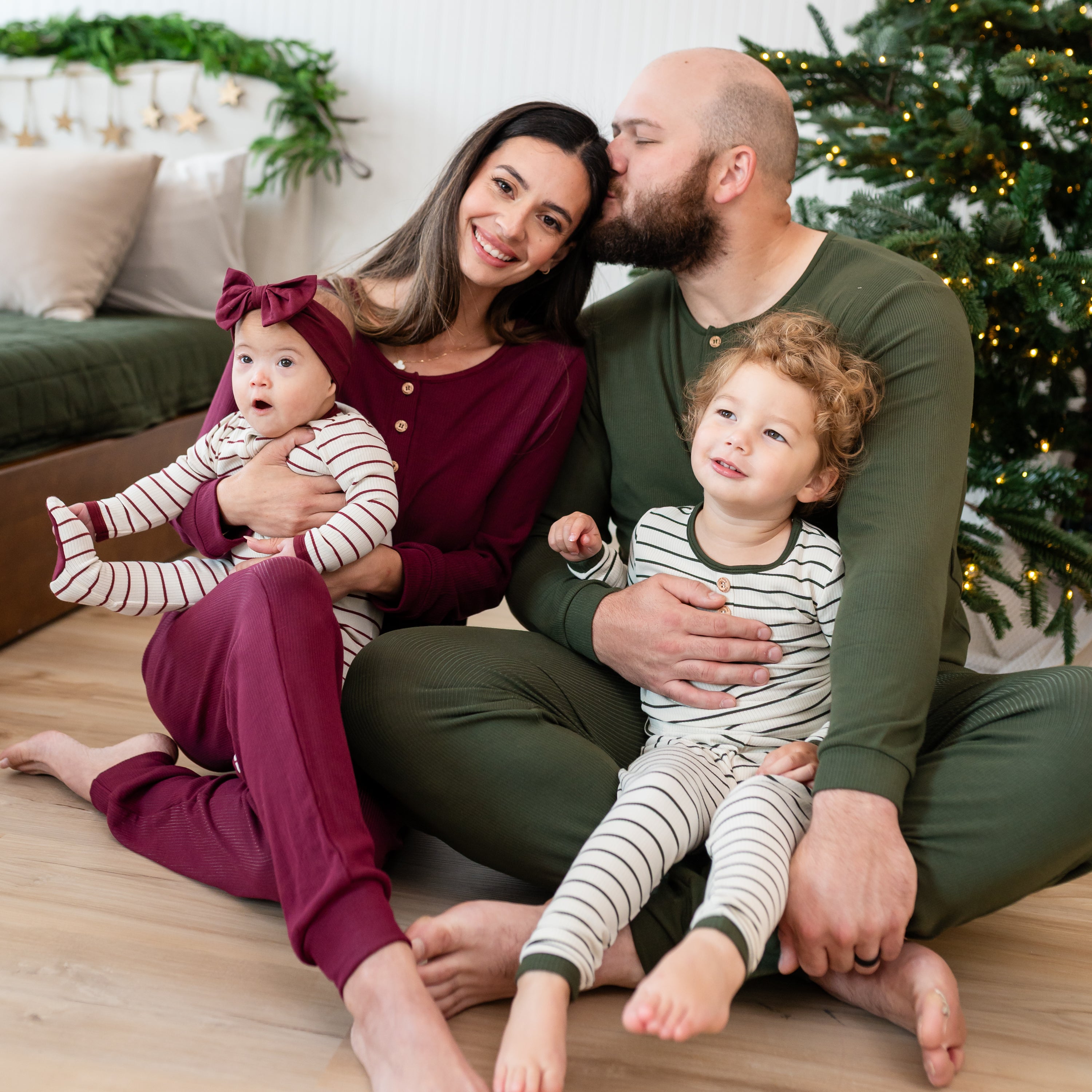 Family of four sitting on the floor in front of a lit Christmas tree wearing corresponding colors. Pictured is the Ribbed Henley Set in Fir Stripe, Men's Ribbed Henley Set in Fir, Women's Ribbed henley Set in Burgundy and ribbed zipper footie in Burgundy stripe