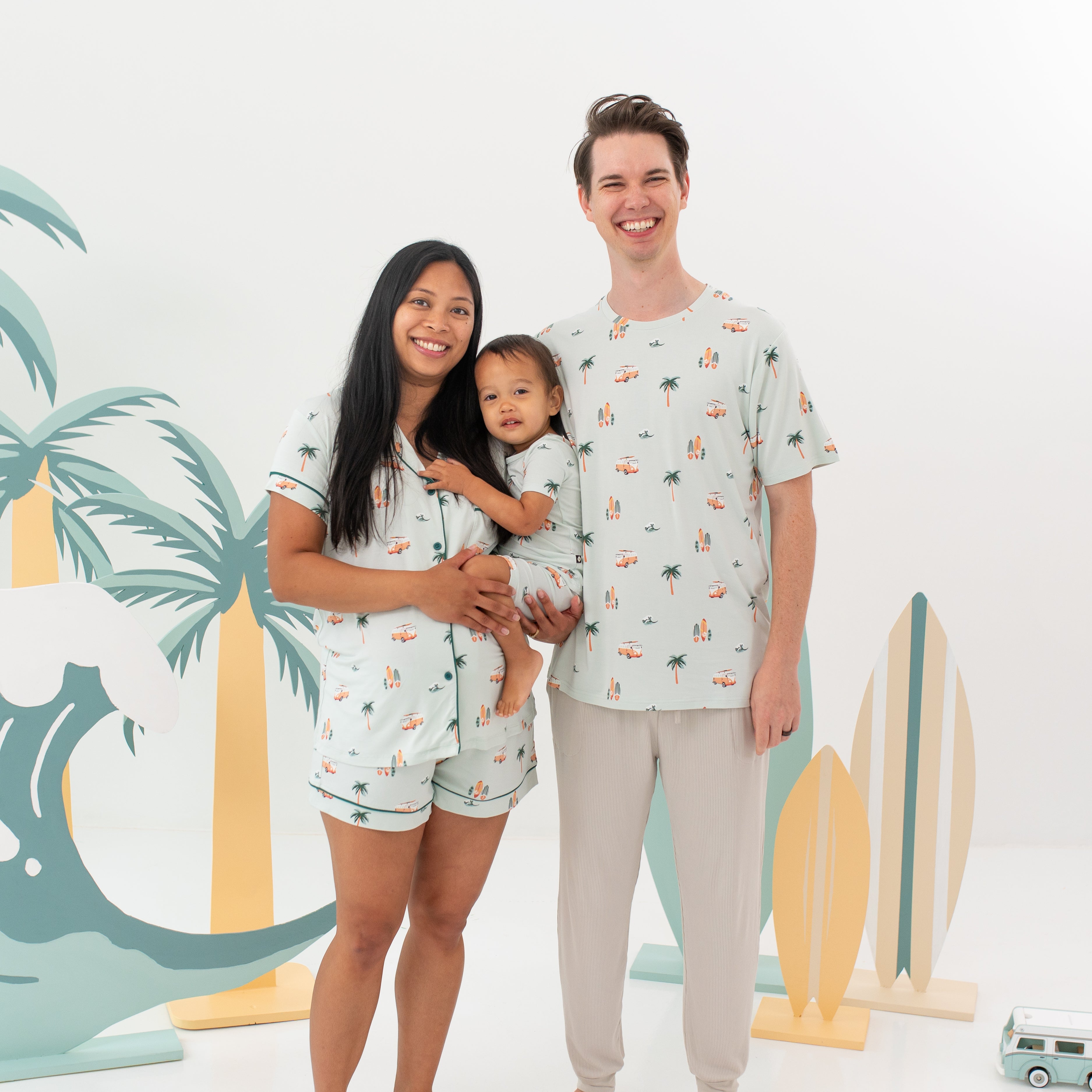 Family matching in Dew Sur standing in front of surf board, palm trees and wave wooden props