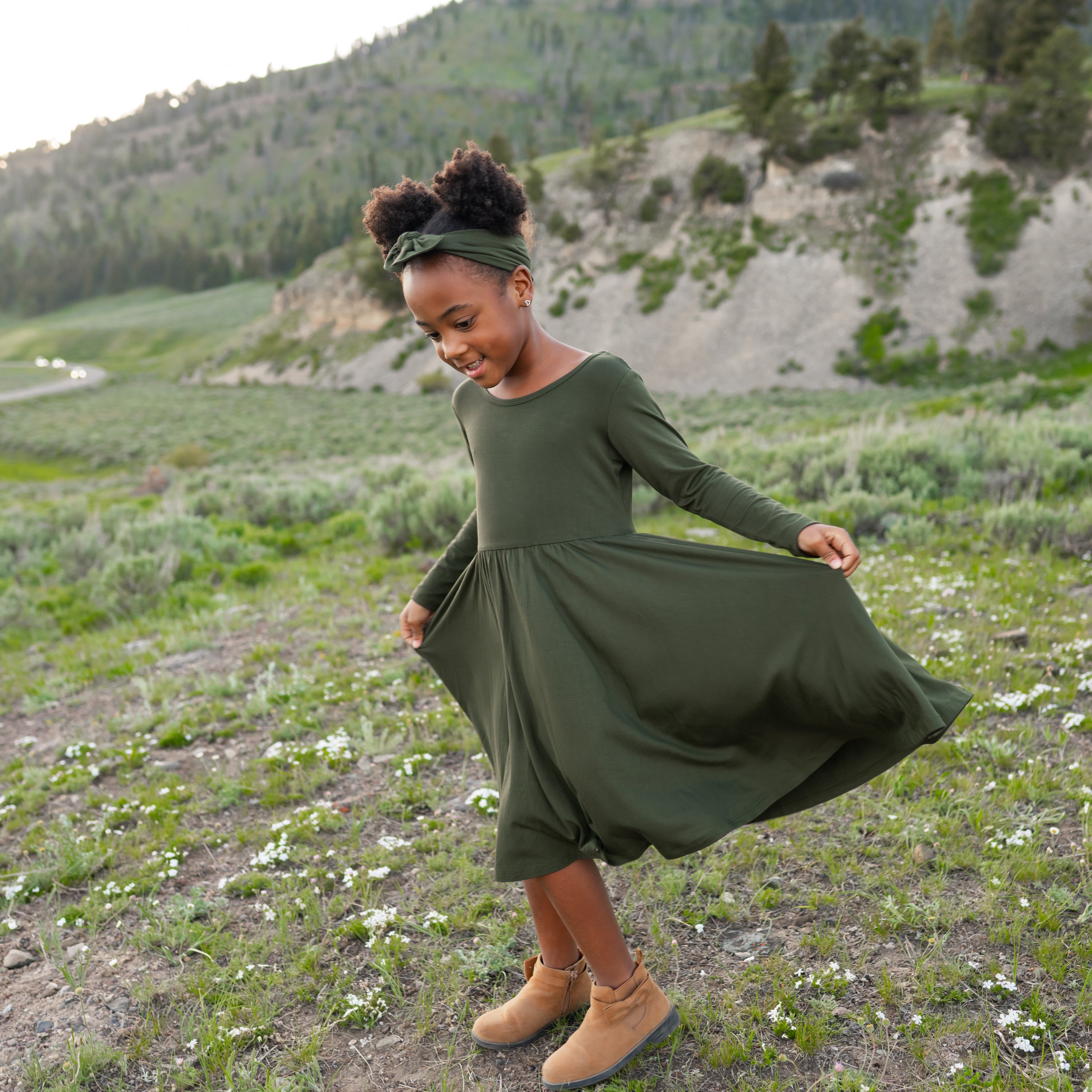Young girl in a green dress standing in a natural landscape with mountains and greenery.