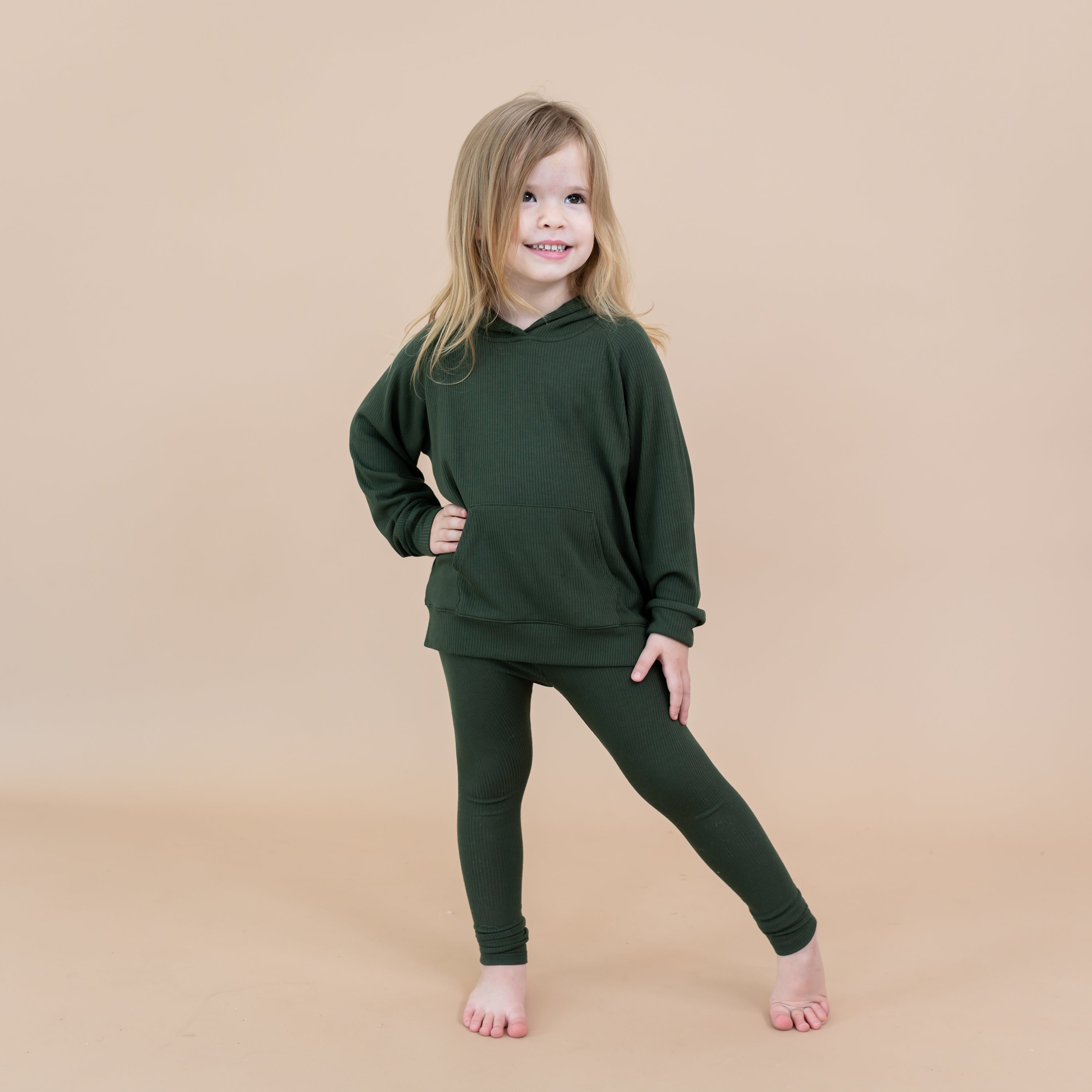 Young girl standing with one leg out to the side wearing the Ribbed Leggings in Fir paired with a matching ribbed hoodie in front of a light beige background