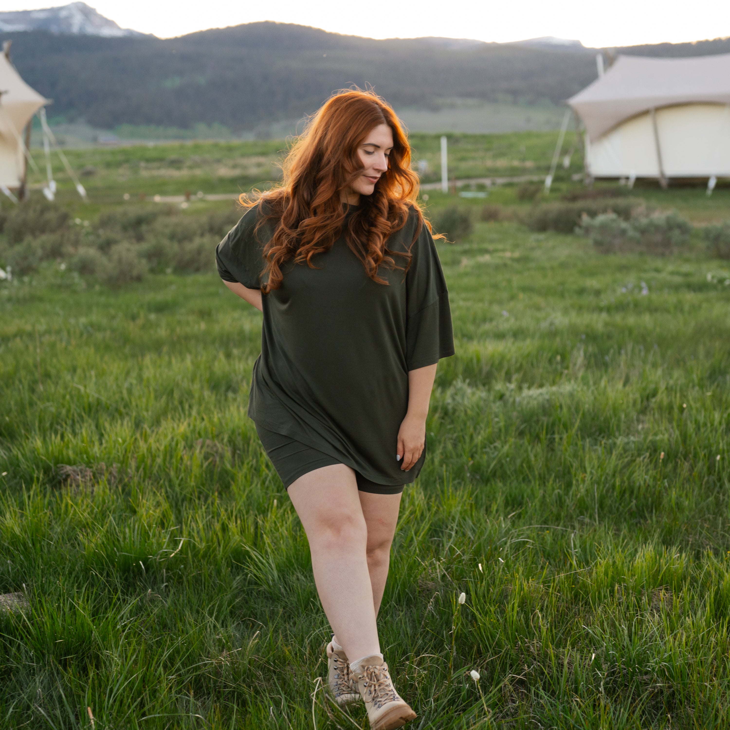 Woman with red hair wearing Women’s Biker Short Set in Fir walking in a grassy field with tents in the background