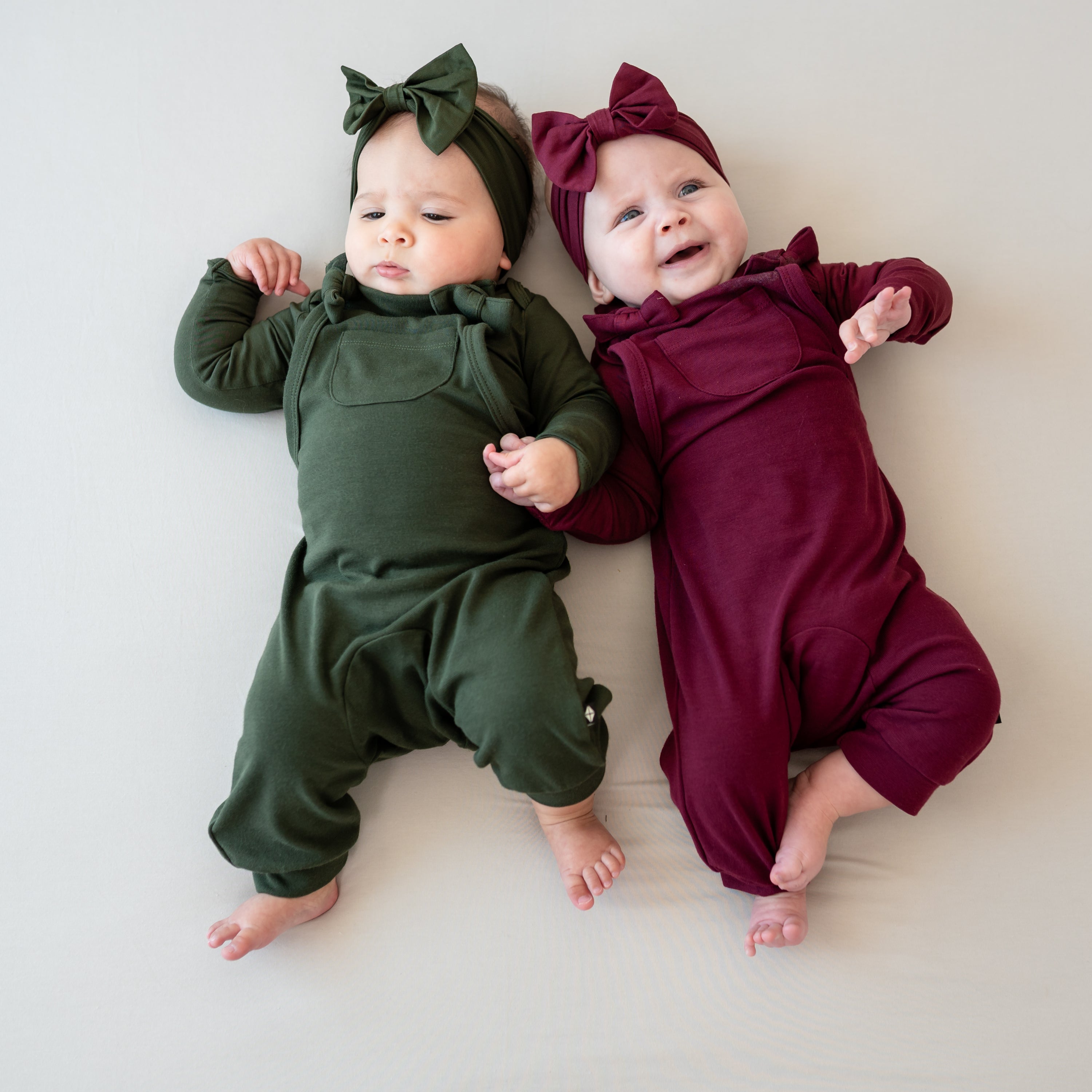 Two infant girls laying side by side wearing the Bamboo Jersey Overalls in Burgundy and Fir and matching long sleeve bodysuits and Bow Headbands