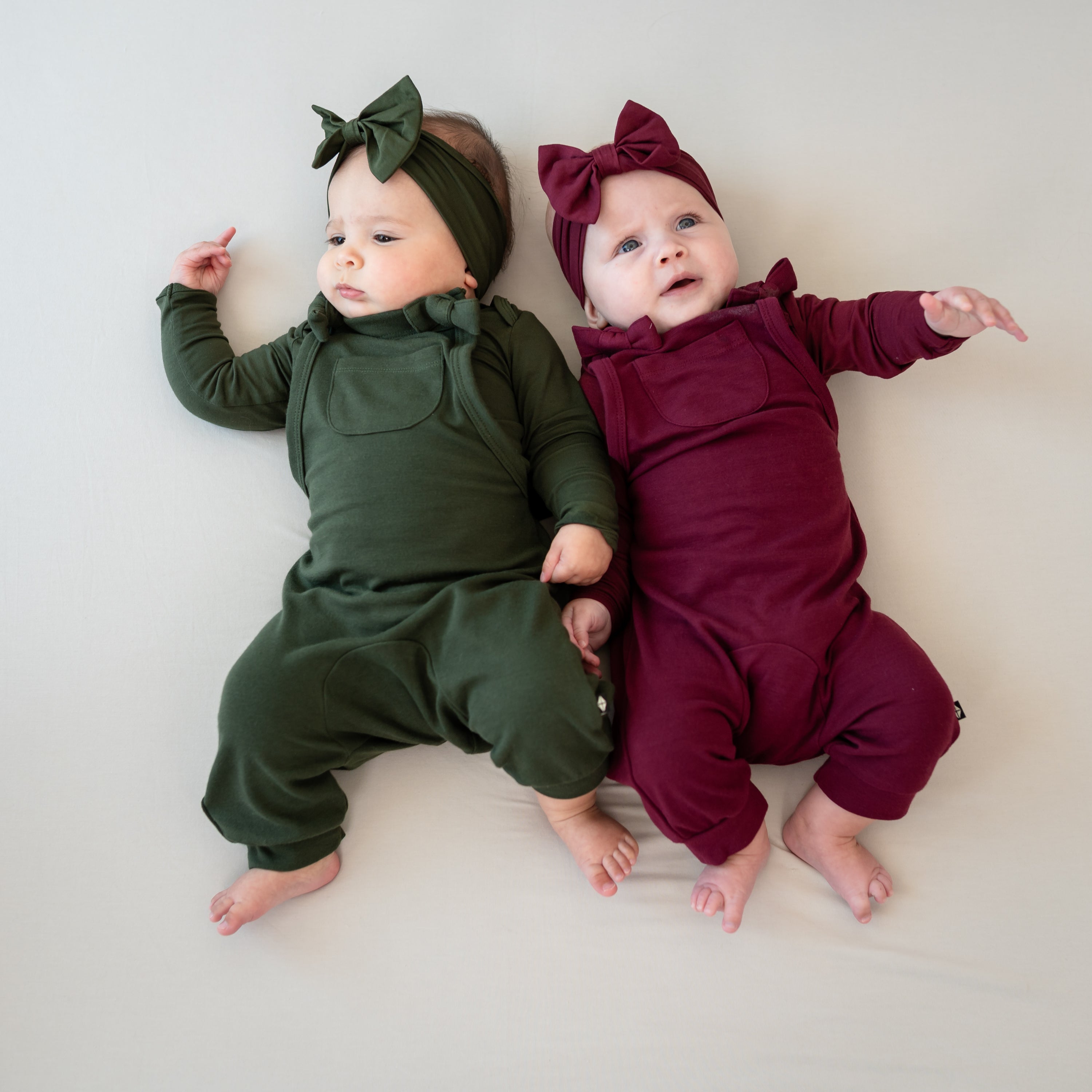Two infant models laying side by side wearing the Bamboo Jersey Overalls in Fir and Burgundy and matching long sleeve bodysuits and bow headbands