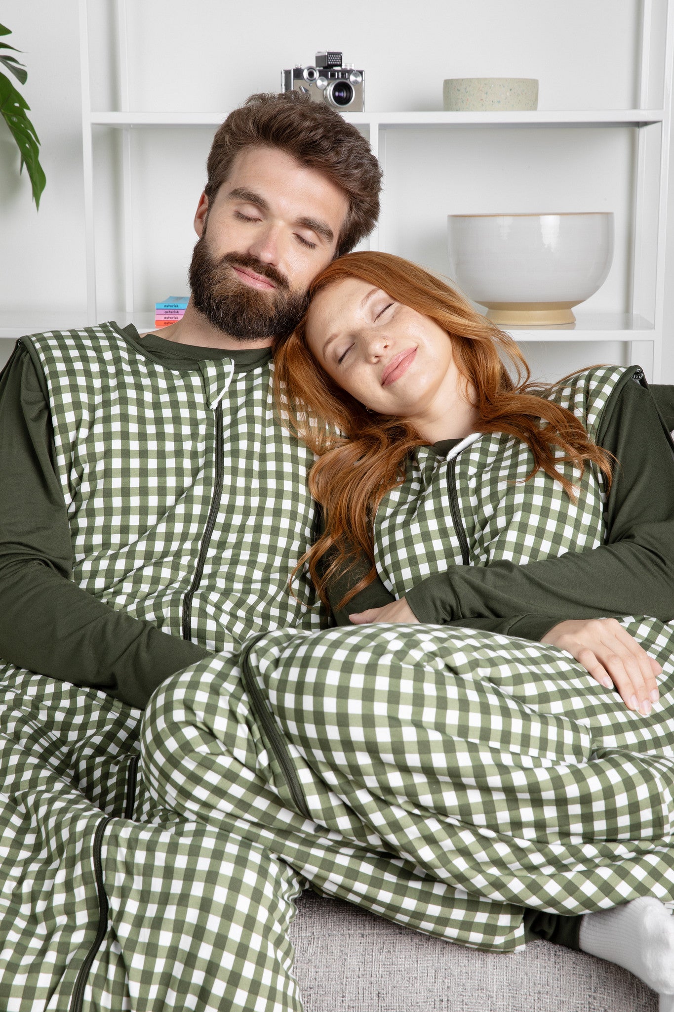 Couple sitting on a couch with their eyes closed both wearing the Adult Sleep Bag Walker in Gingham Fir