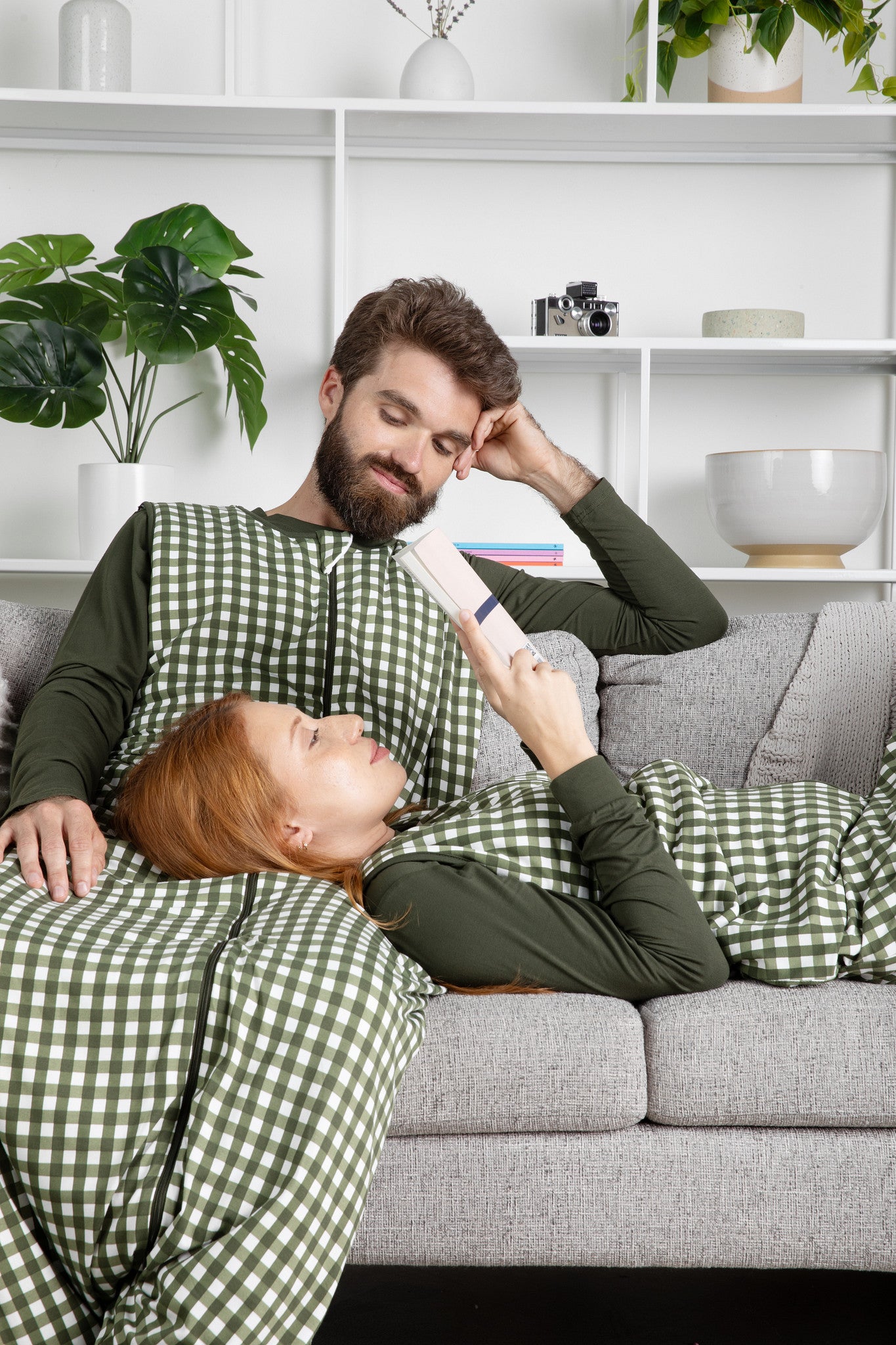 Couple sitting on a couch with the wife laying on the husbands lap reading a book both wearing the Adult Sleep Bag Walker in Gingham Fir