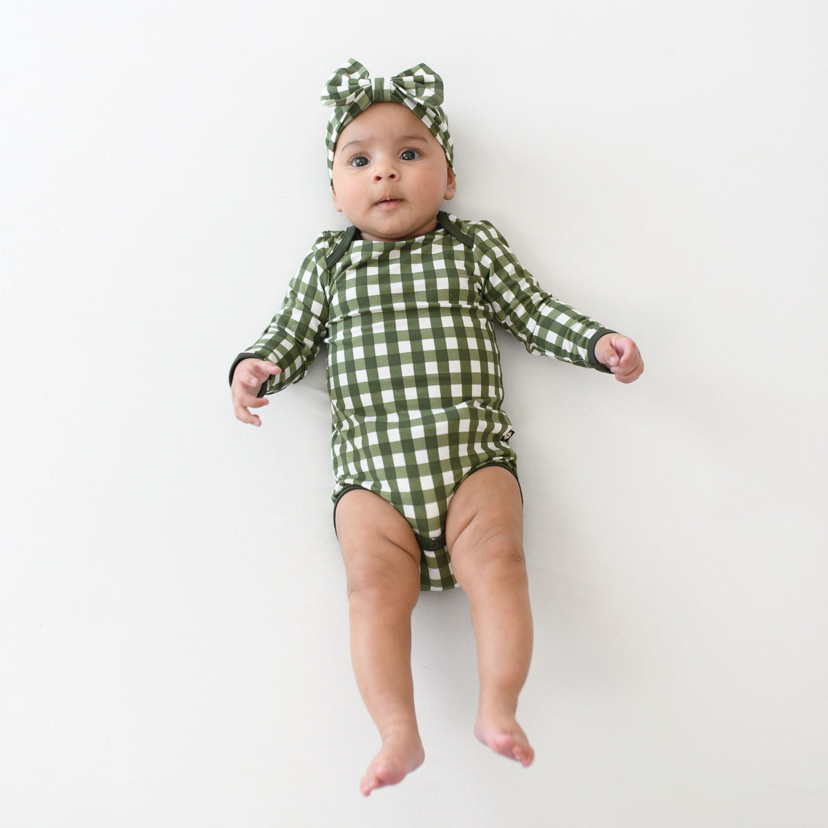 Infant laying down wearing the Long Sleeve Bodysuit in Gingham Fir with matching bow headband