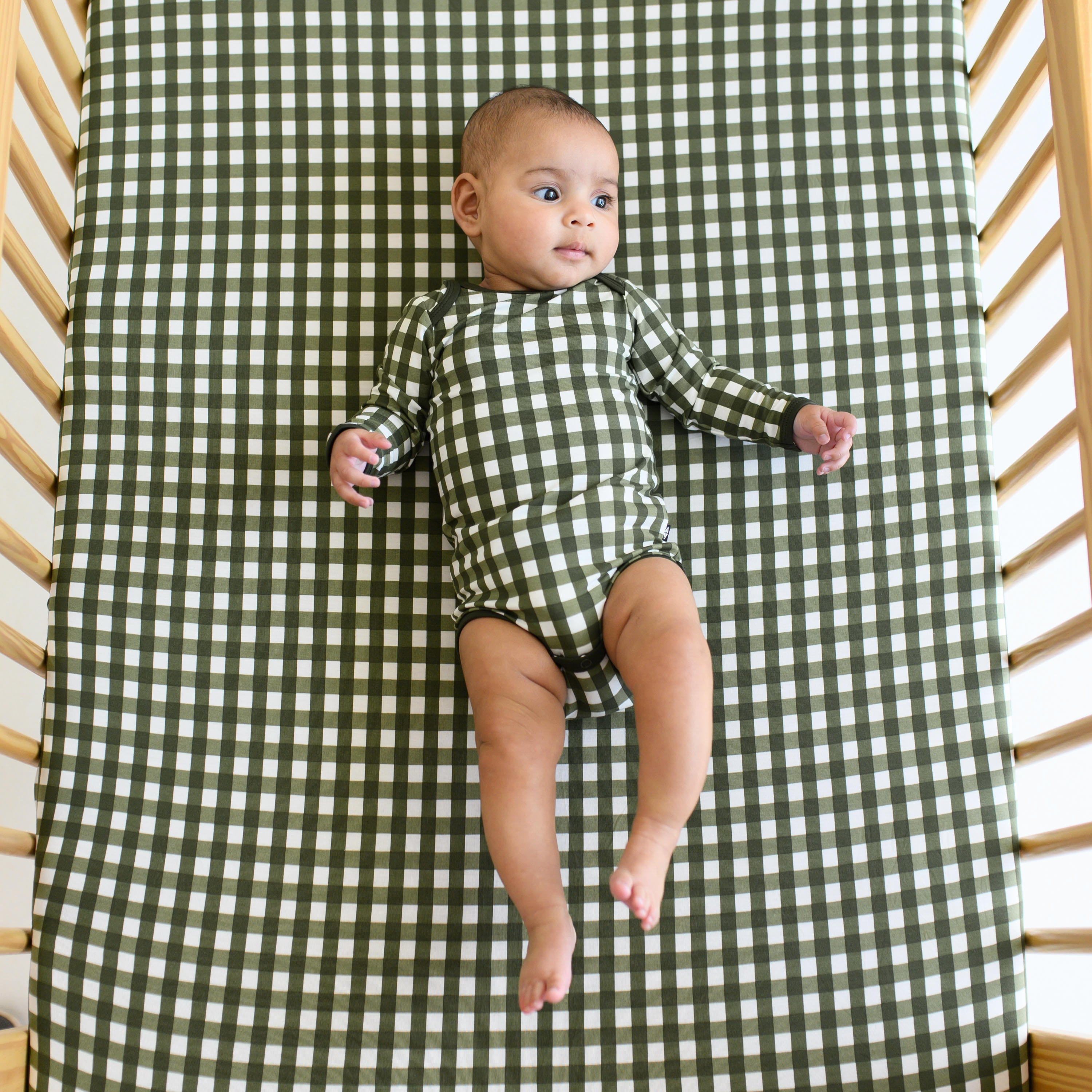 Infant laying in a crib on top of the Crib Sheet in Gingham Fir wearing a matching long sleeve bodysuit