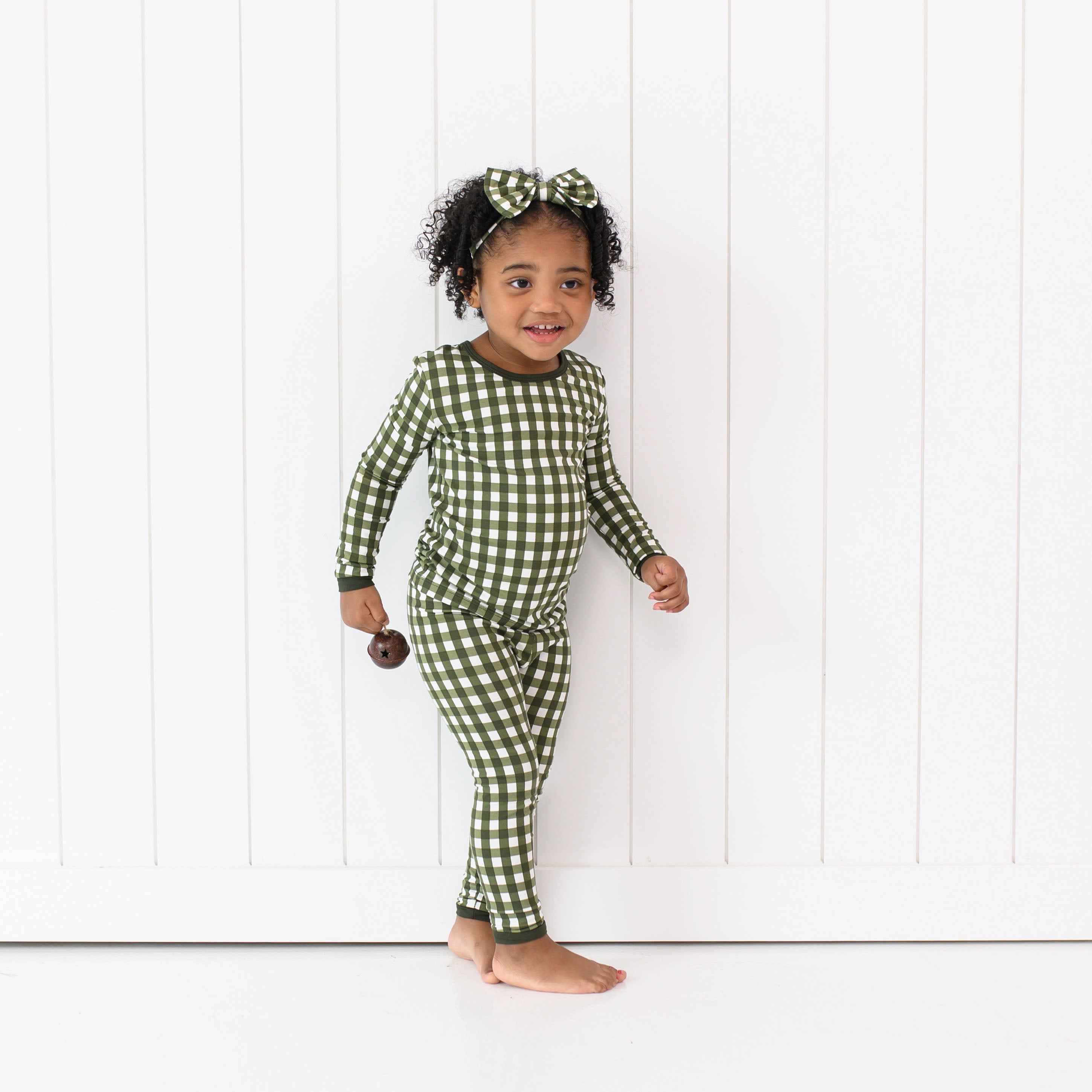 Young girls standing in front of a white paneled wall wearing the Long Sleeve Pajamas in Gingham Fir with matching bow headband