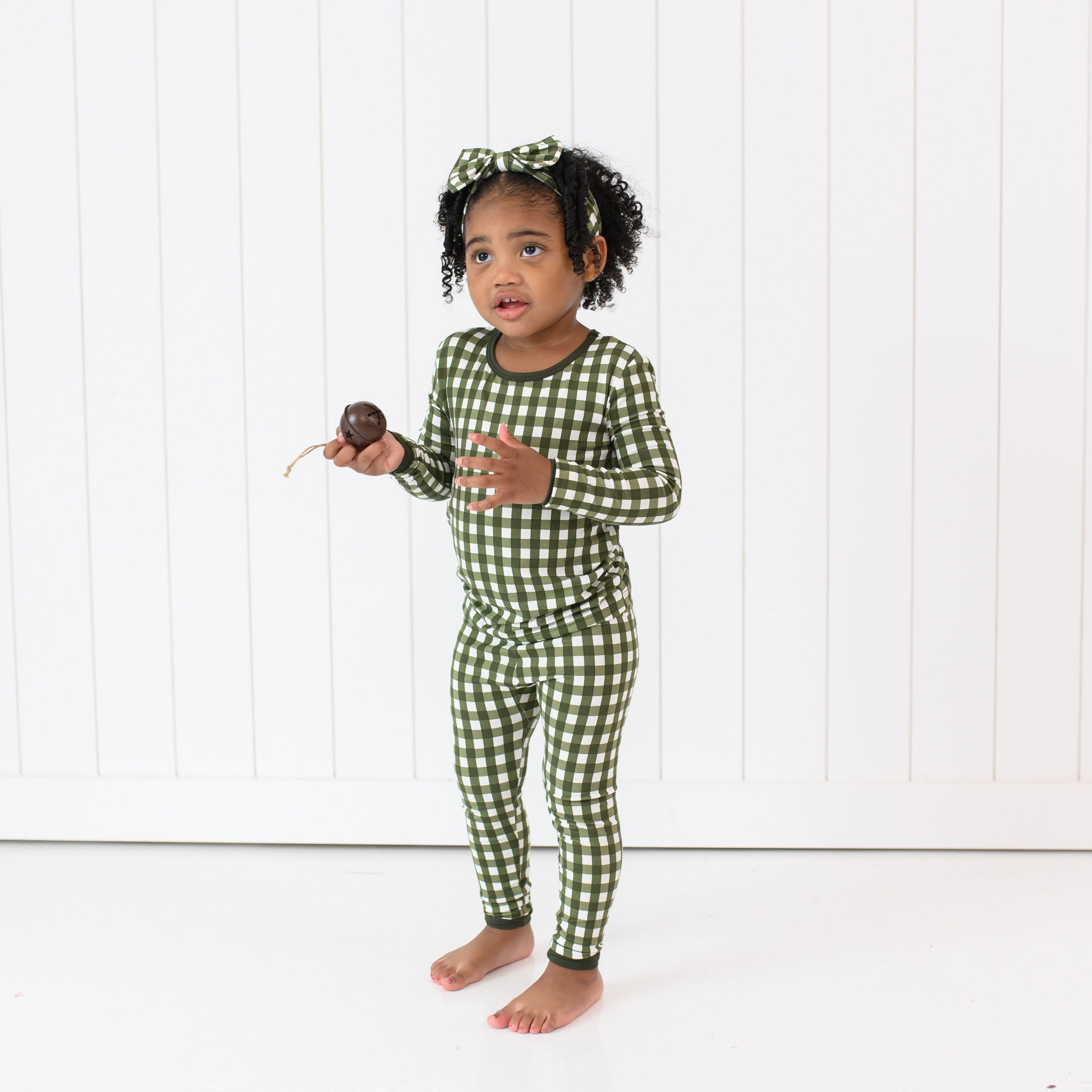 Toddler standing in front of a white paneled wall wearing Kyte Long Sleeve Pajamas in Gingham Fir