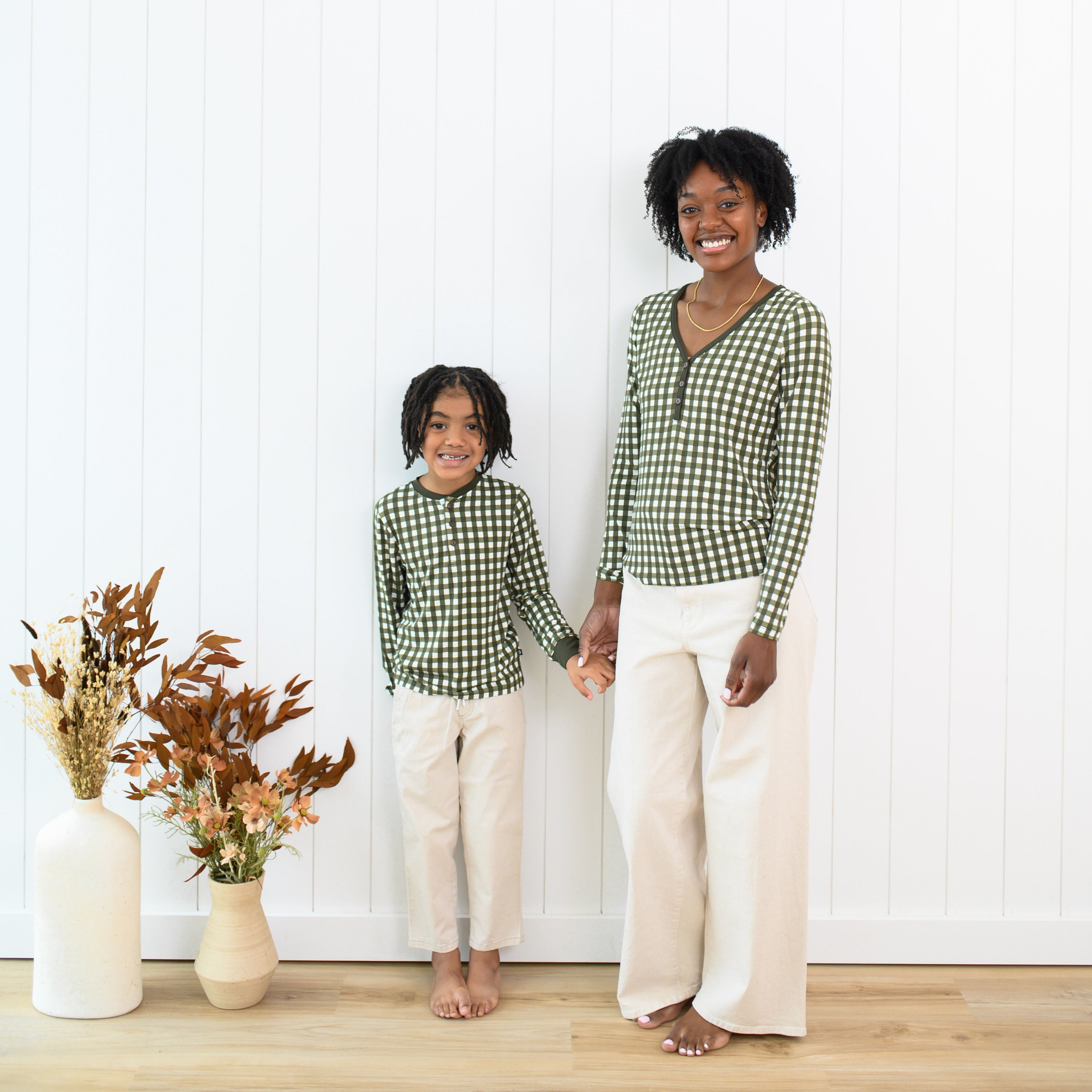 Mother and daughter holding hands standing in front of a white paneled wall wearing the Women's Long Sleeve Henley Tee in Gingham Fir