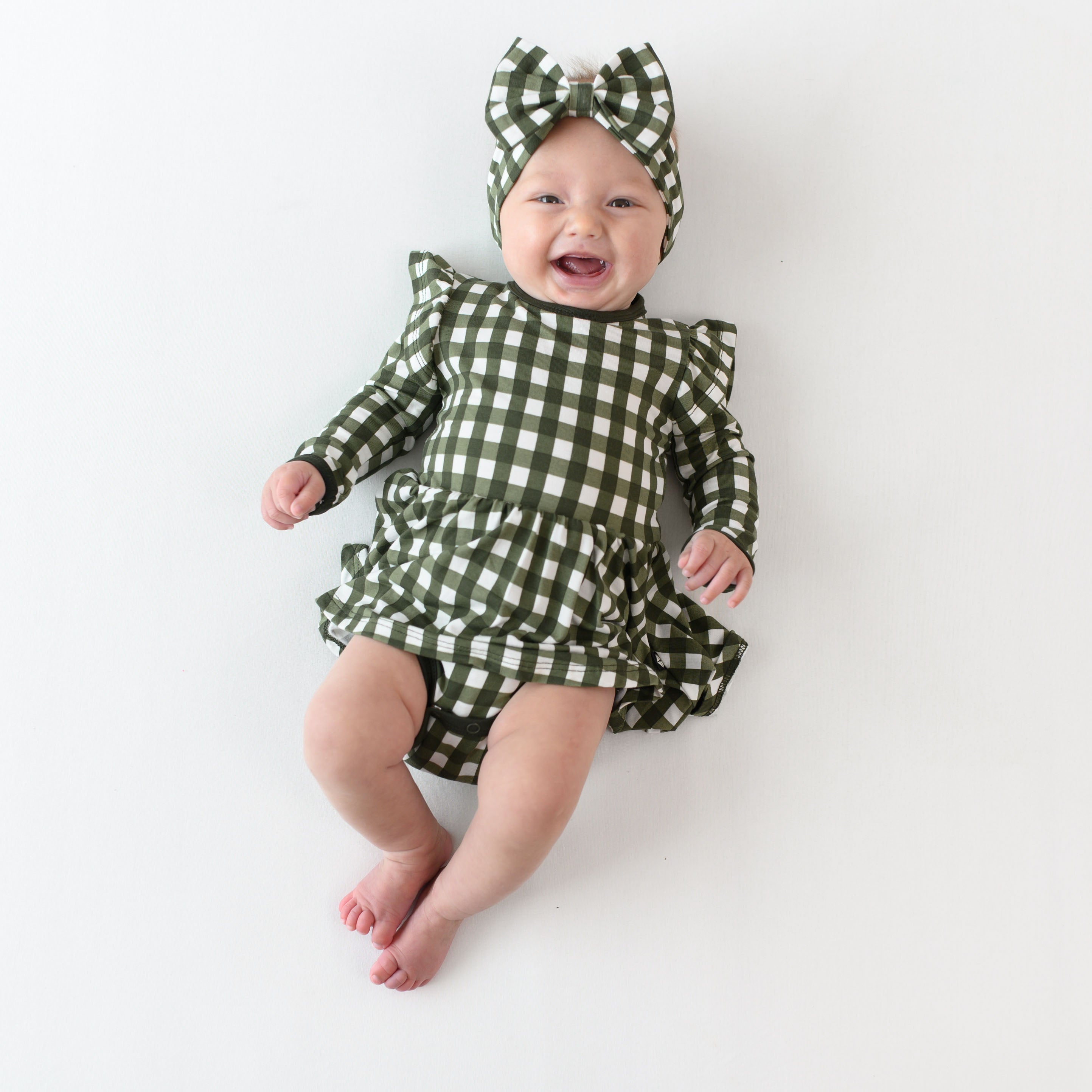 Smiling infant laying down wearing the Long Sleeve Twirl Bodysuit Dress in Gingham Fir and matching bow headband