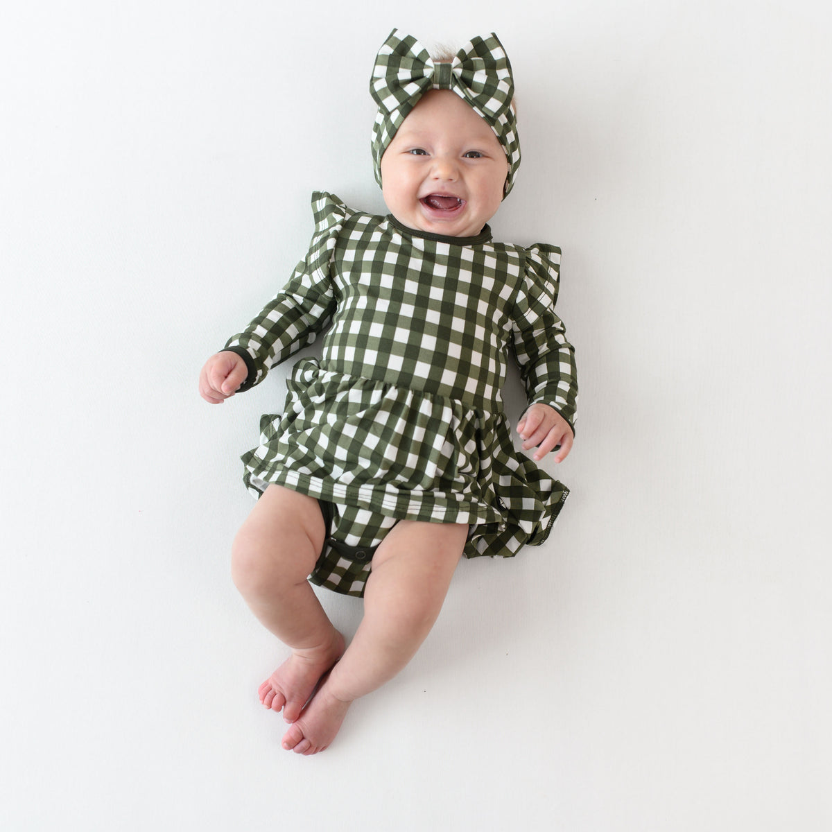 Smiling infant laying down wearing the Long Sleeve Twirl Bodysuit Dress in Gingham Fir and matching bow headband