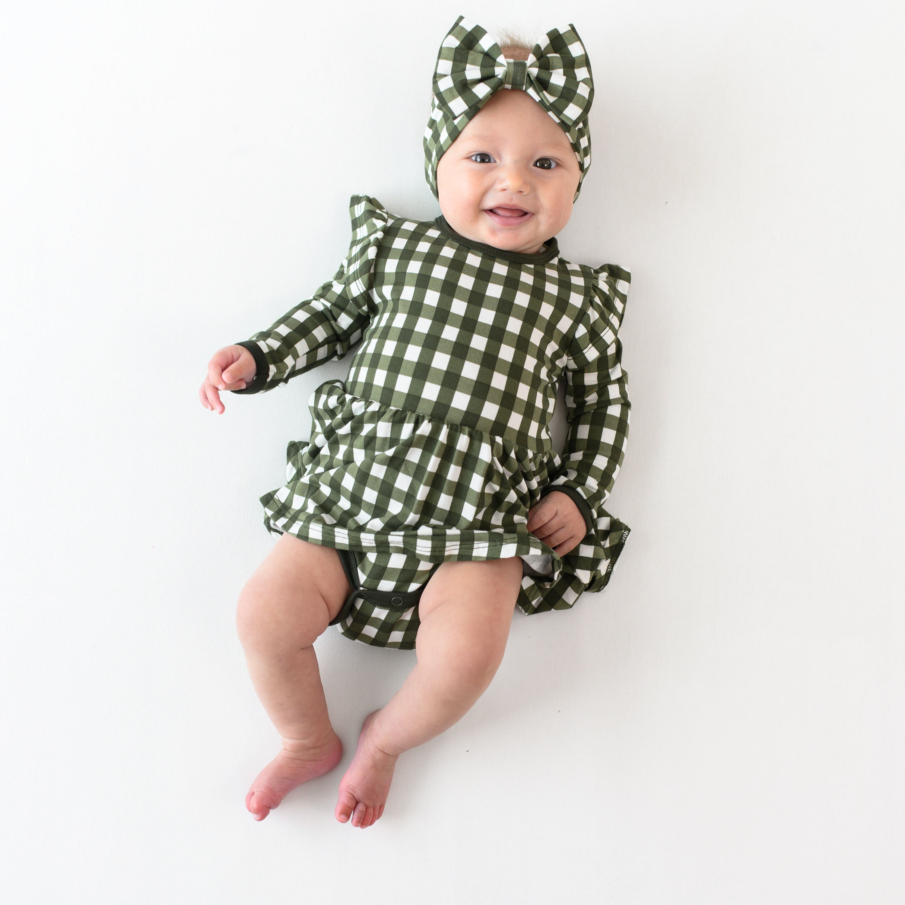 Infant girl laying down wearing the Long Sleeve Twirl Bodysuit Dress in Gingham Fir with matching bow headband