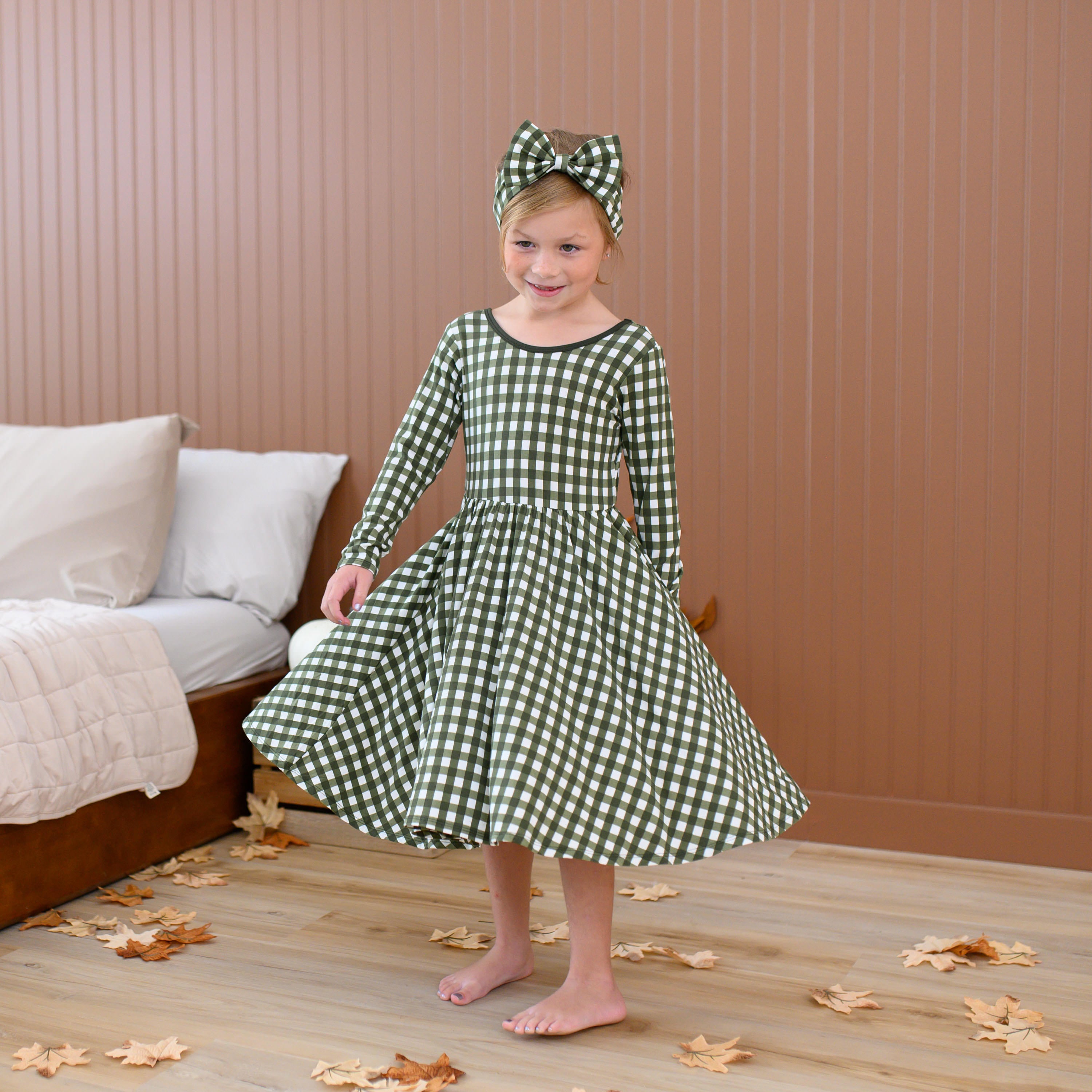 Girl twirling beside a bed wearing the Long Sleeve Twirl Dress in Gingham Fir and matching bow headband