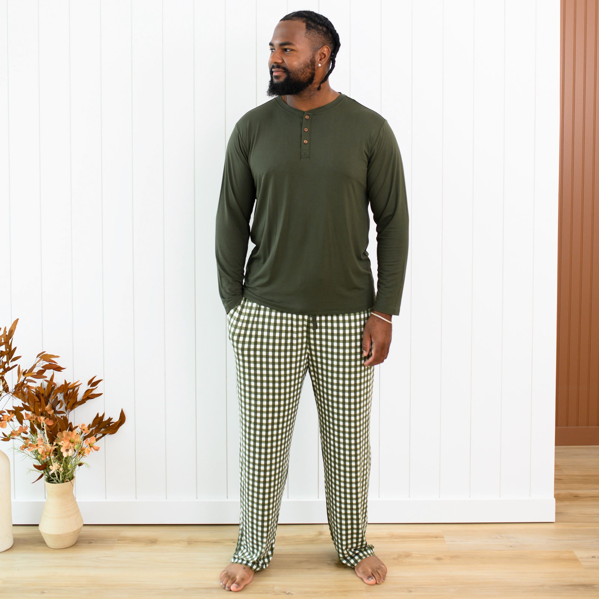 Male model standing in front of a white paneled wall wearing the Men's Lounge Pants in Gingham Fir with Men's Fir Henley top