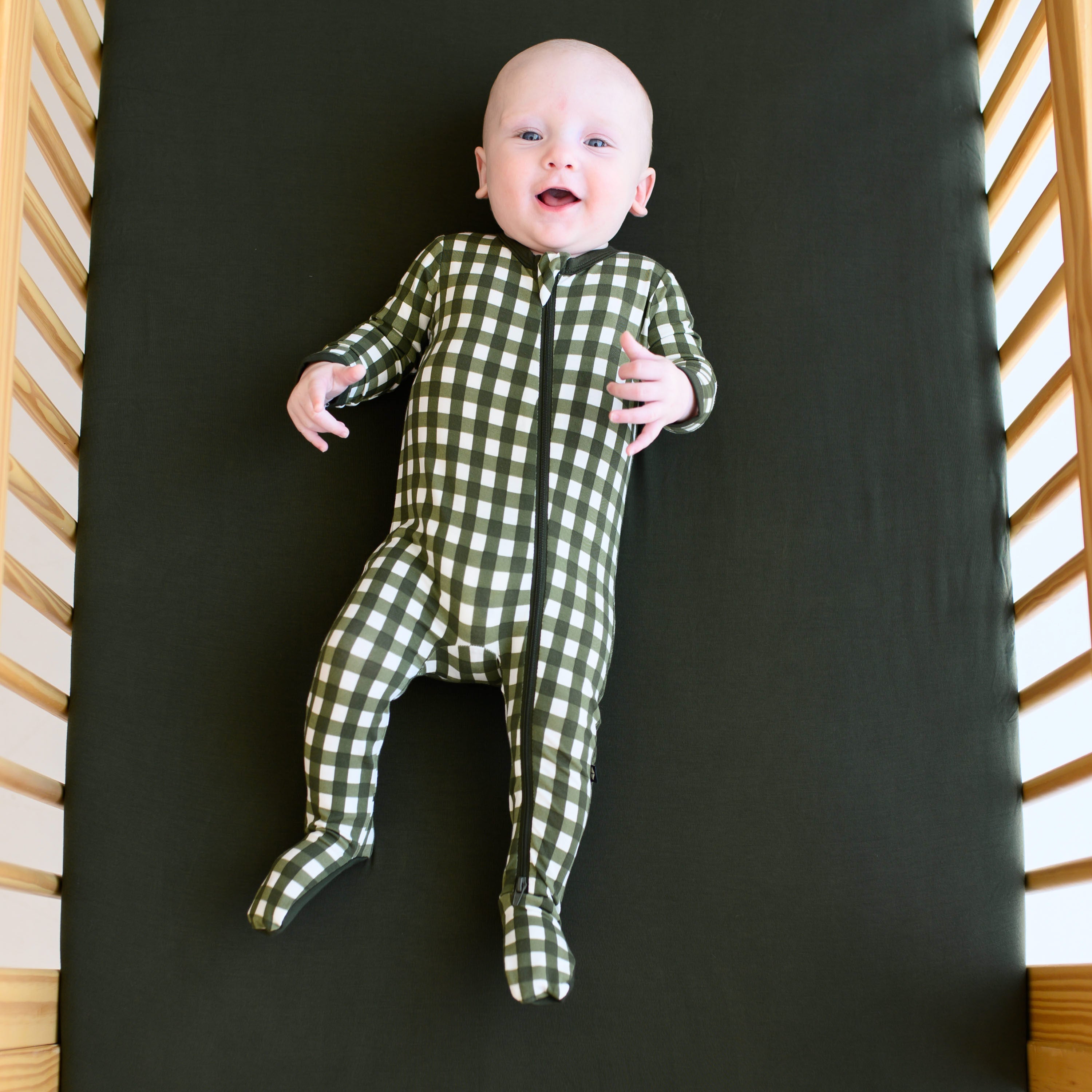 Smiling infant wearing the Zippered Footie in Gingham Fir laying in a crib on a Fir Crib Sheet