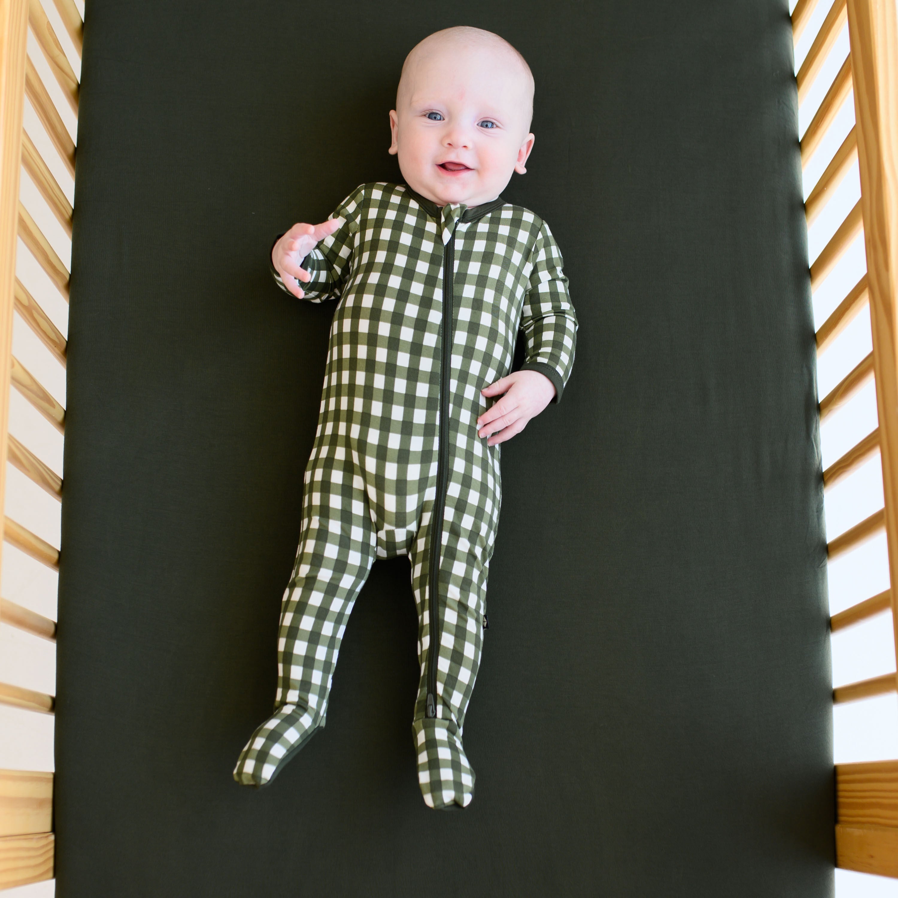 Infant laying in a crib on top of a Fir Crib Sheet wearing the Zippered Footie in Gingham Fir