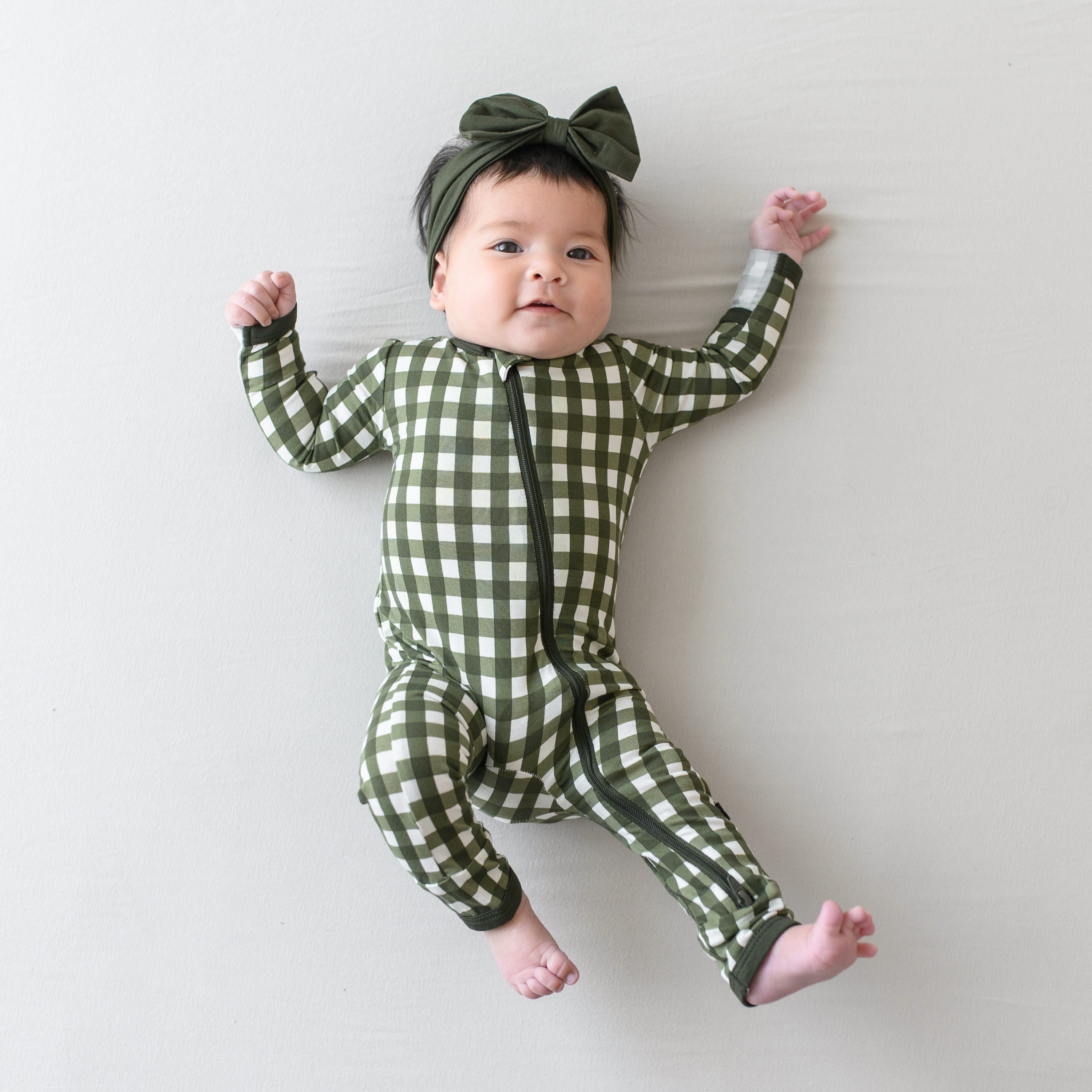 Infant girl laying down wearing the Zippered Romper in Gingham Fir with Fir Bow Headband