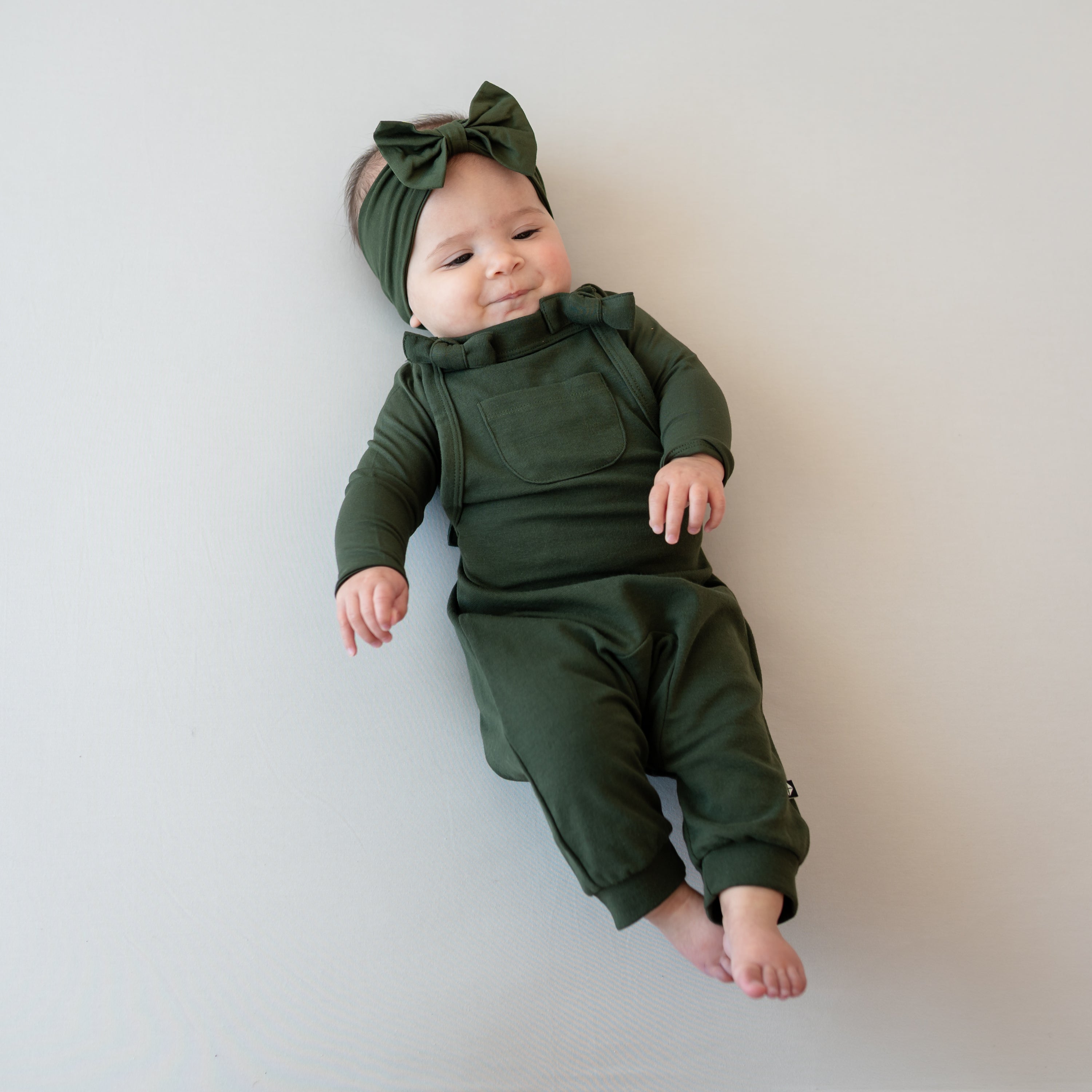 Infant girl laying down on a light neutral background wearing the Bamboo Jersey Overall in Fir with long sleeve matching body suit and bow headband as an accessory
