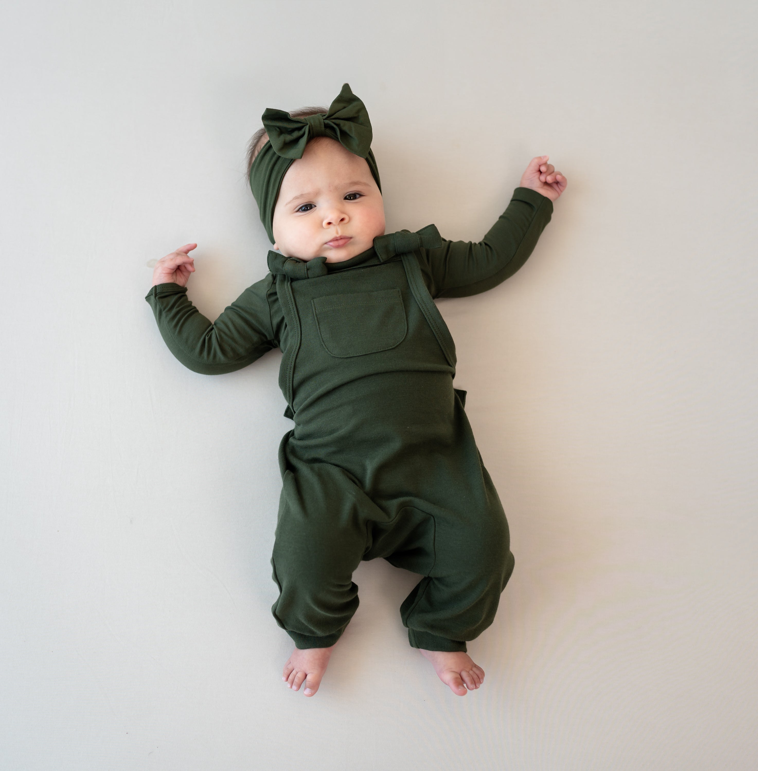 Infant girl laying down wearing the Bamboo Jersey Overall in Fir with matching long sleeve bodysuit and Bow headband
