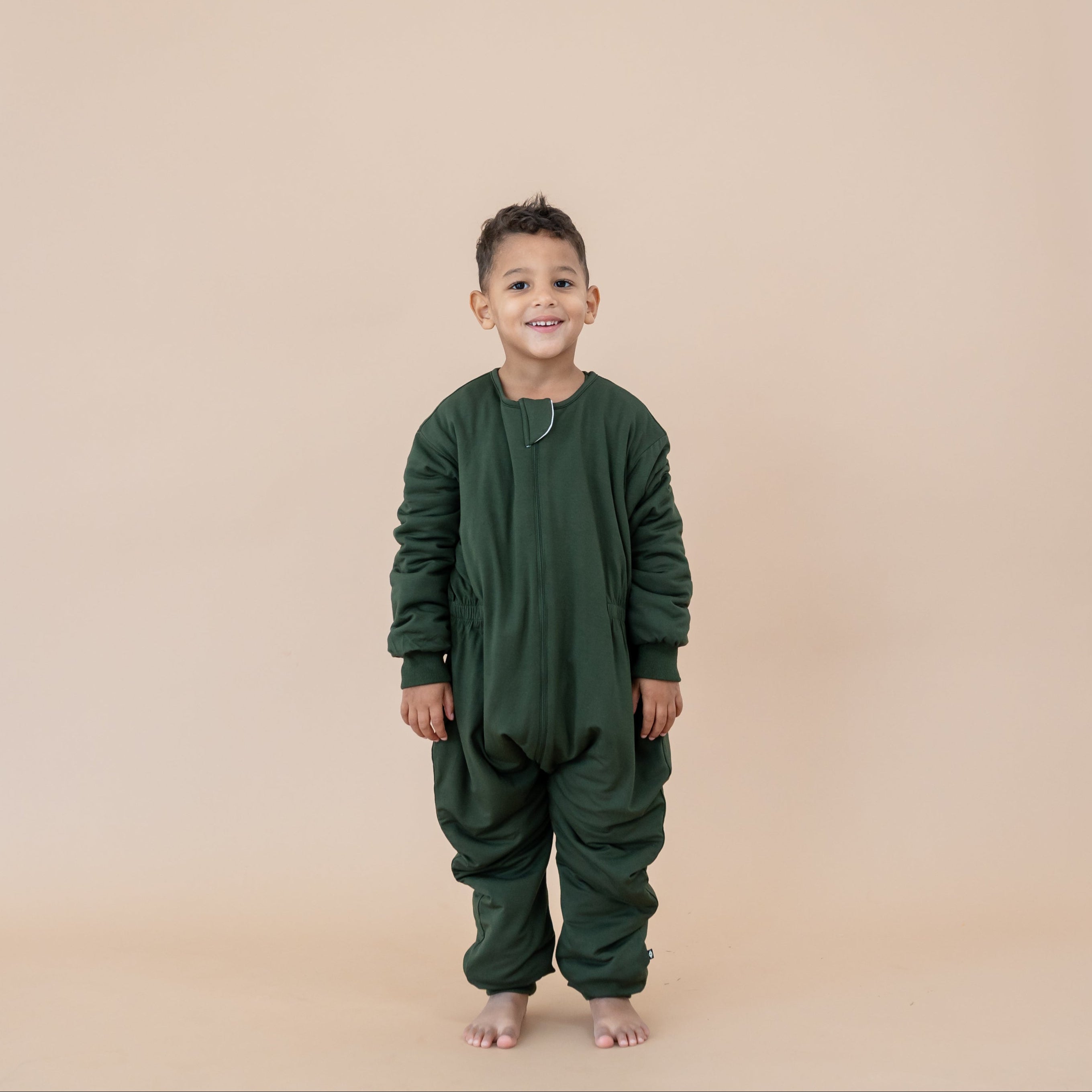 Young boy standing with his hands at his side wearing the Cozy Playsuit in Fir in front of a light taupe backdrop
