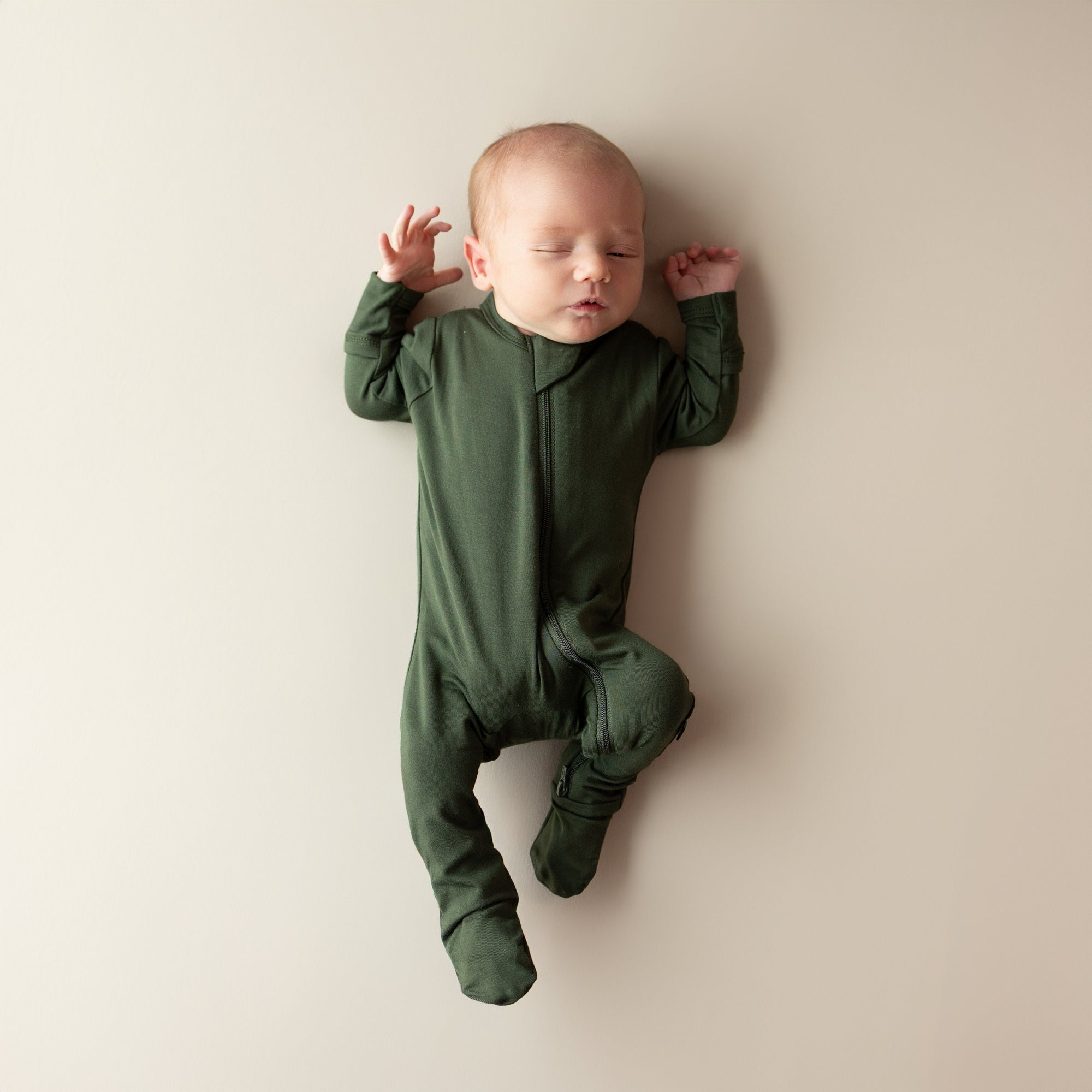 Baby in Kyte Baby bamboo Zippered Footie in Fir lying on a beige surface