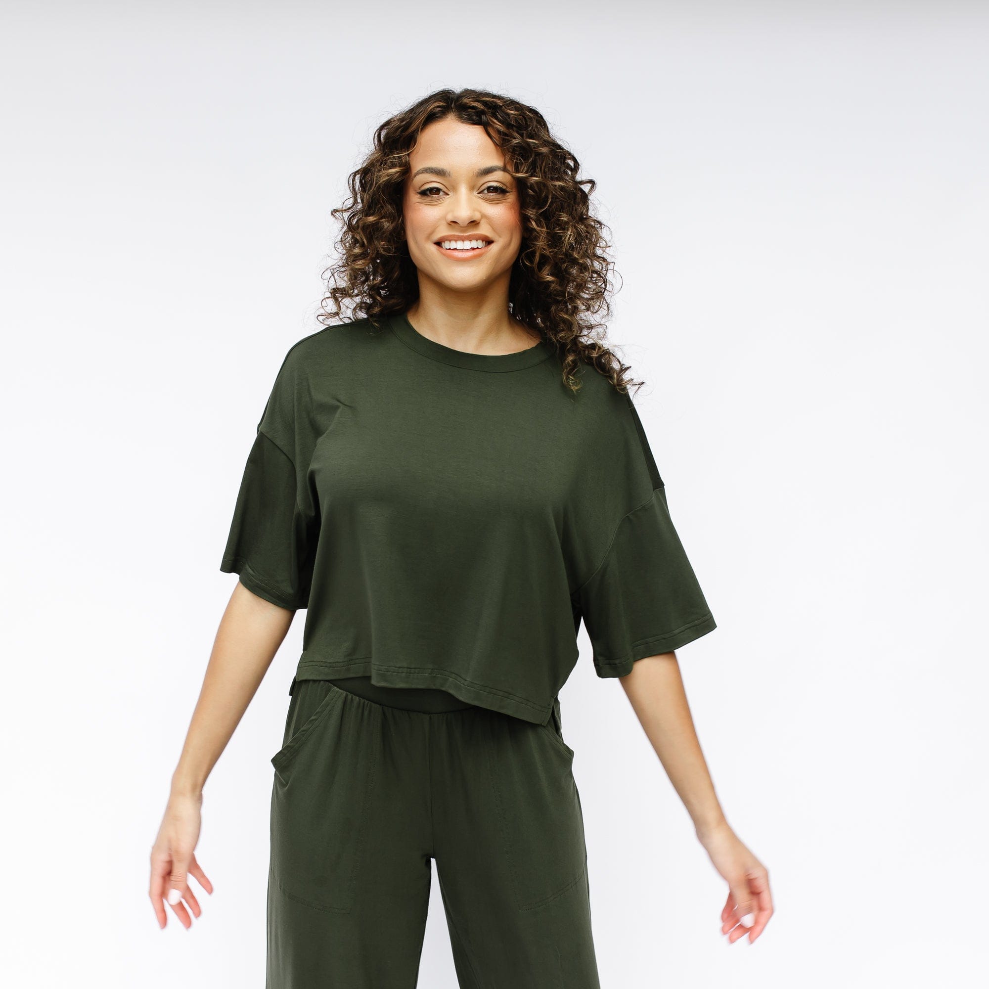 Smiling female model standing in front of a white wall wearing the Women's Short Sleeve Tee in Fir with matching wide leg pants