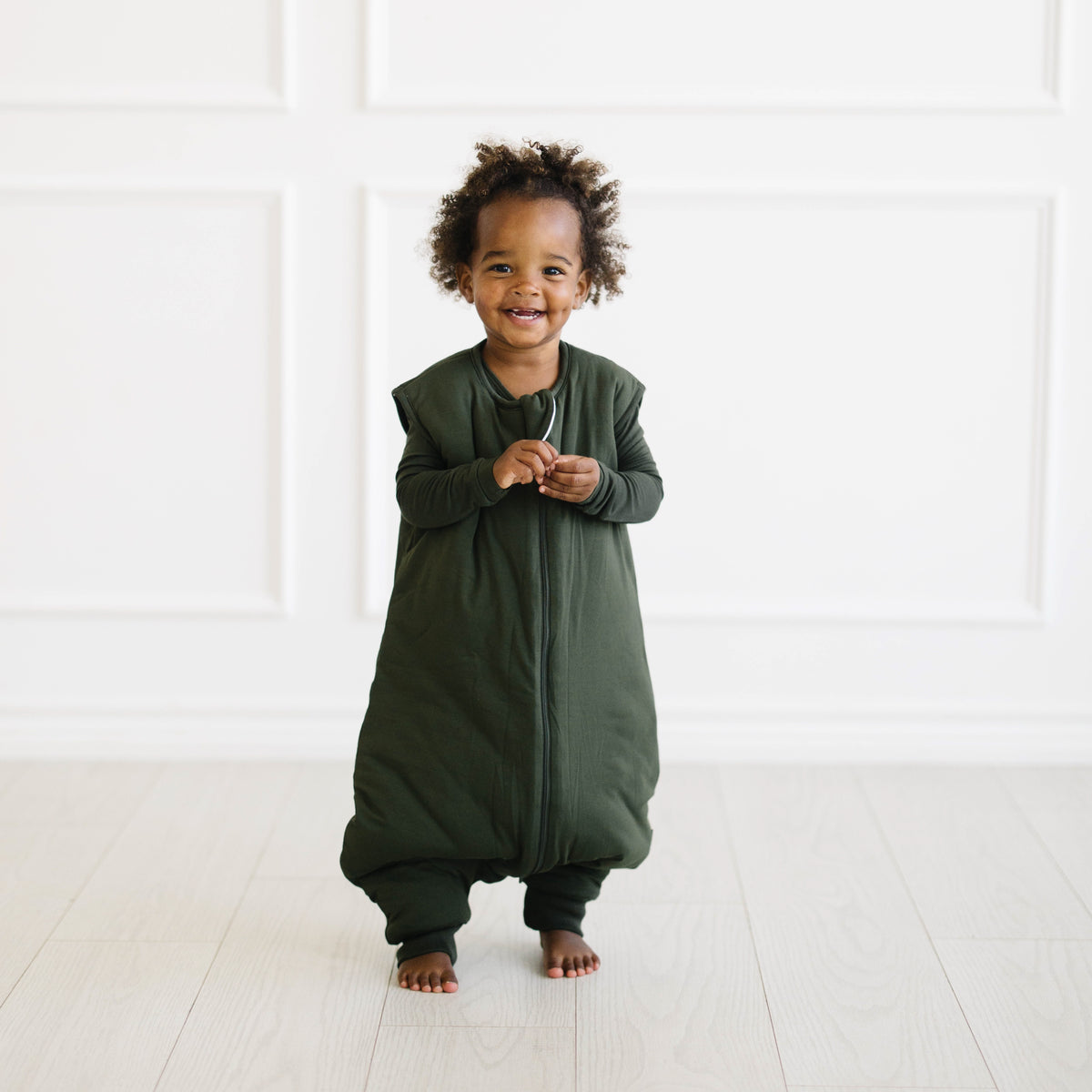 Child wearing Sleep Bag Walker in Fir 2.5 standing on a white floor with a white wall background