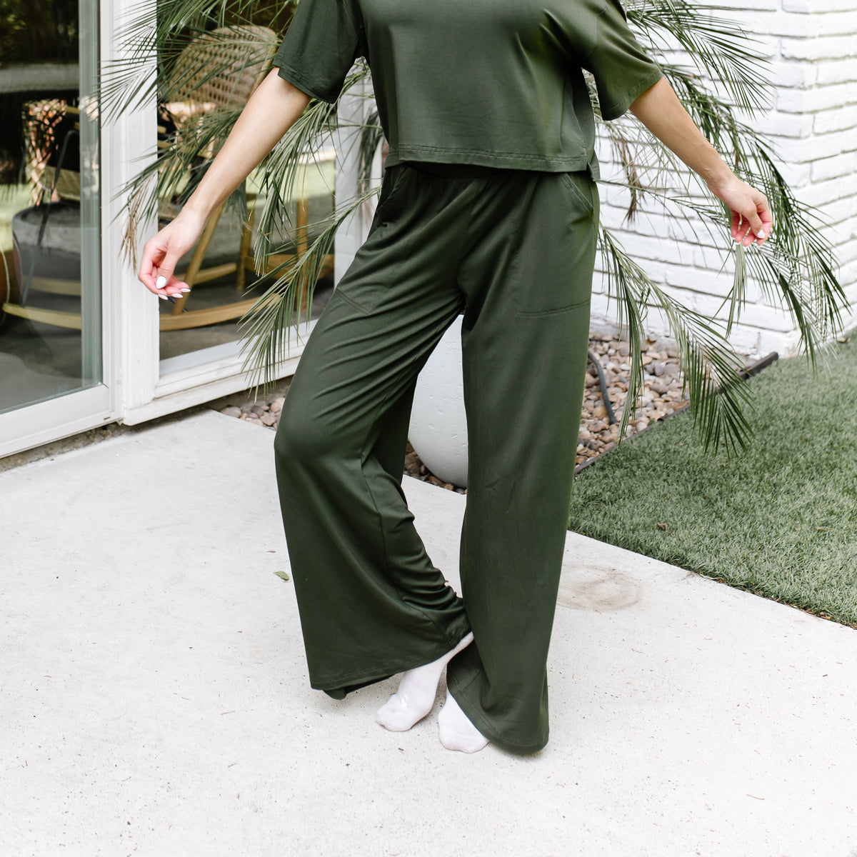 Female model standing on a sidewalk in front of a white brick house wearing the Women's Wide Leg Pant in Fir