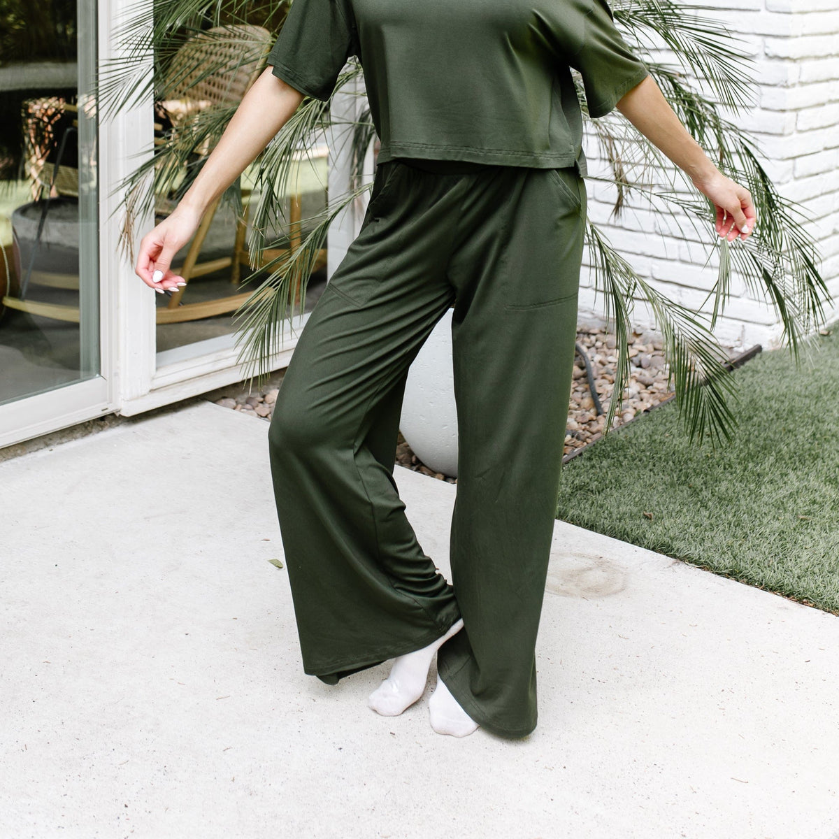 Female model standing on a sidewalk in front of a white brick house wearing the Women's Wide Leg Pant in Fir