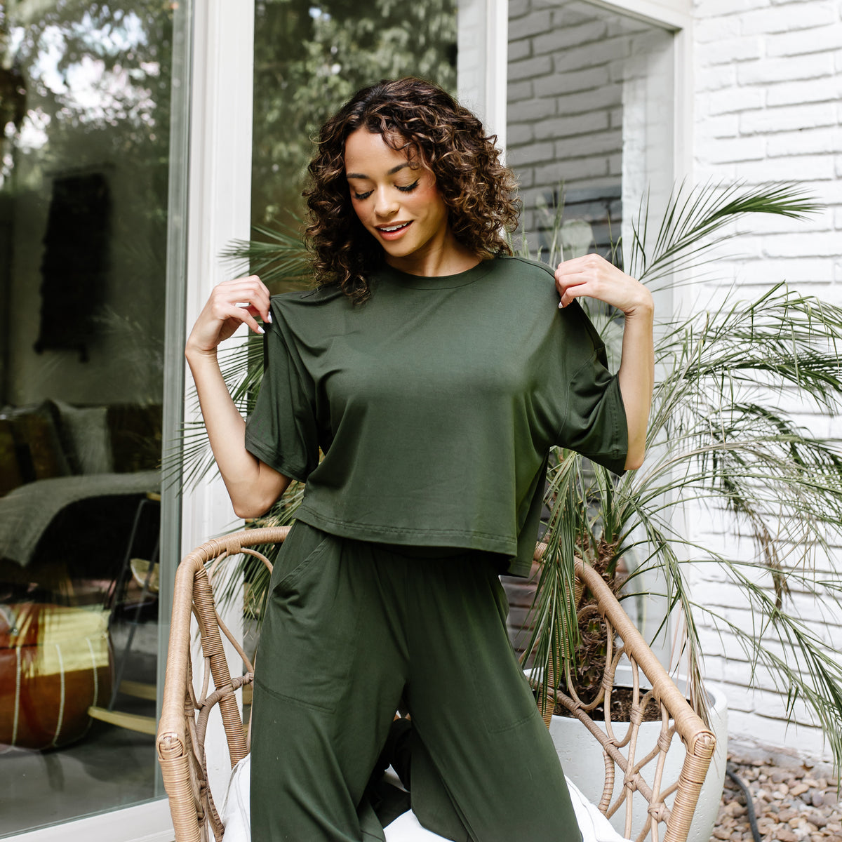 Female model standing in front of a white brick house wearing the Women's Short Sleeve Tee in Fir