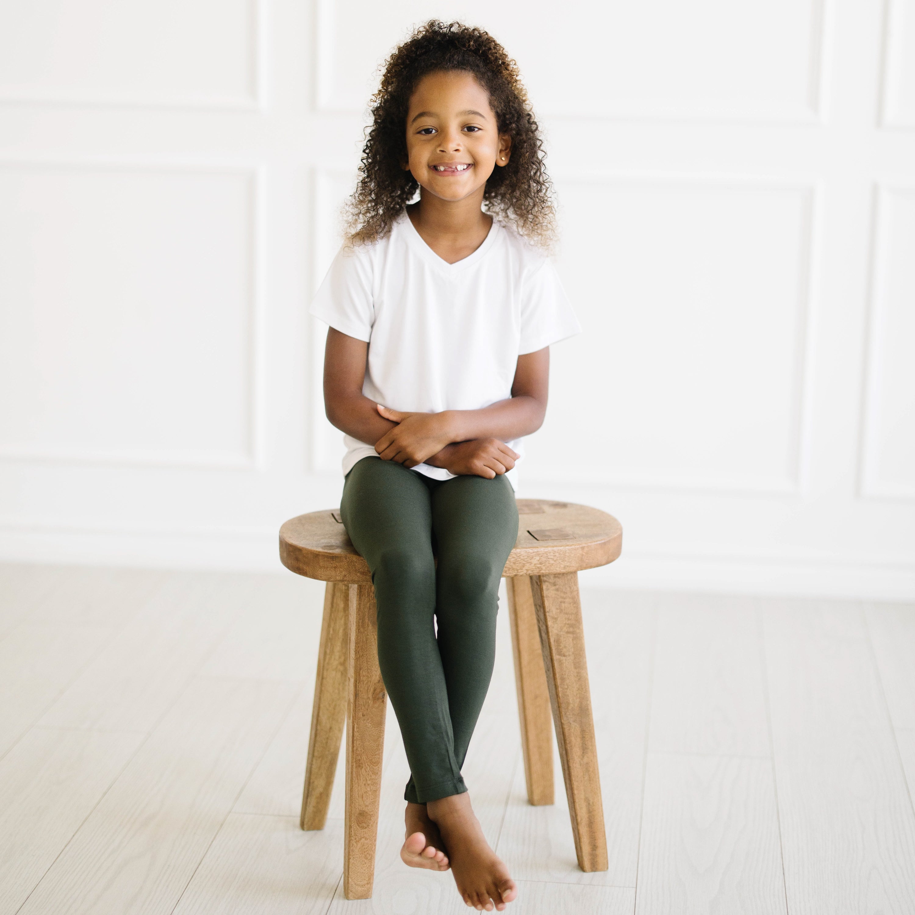 Child sitting on a wooden stool wearing a white shirt and green leggings in a bright room.