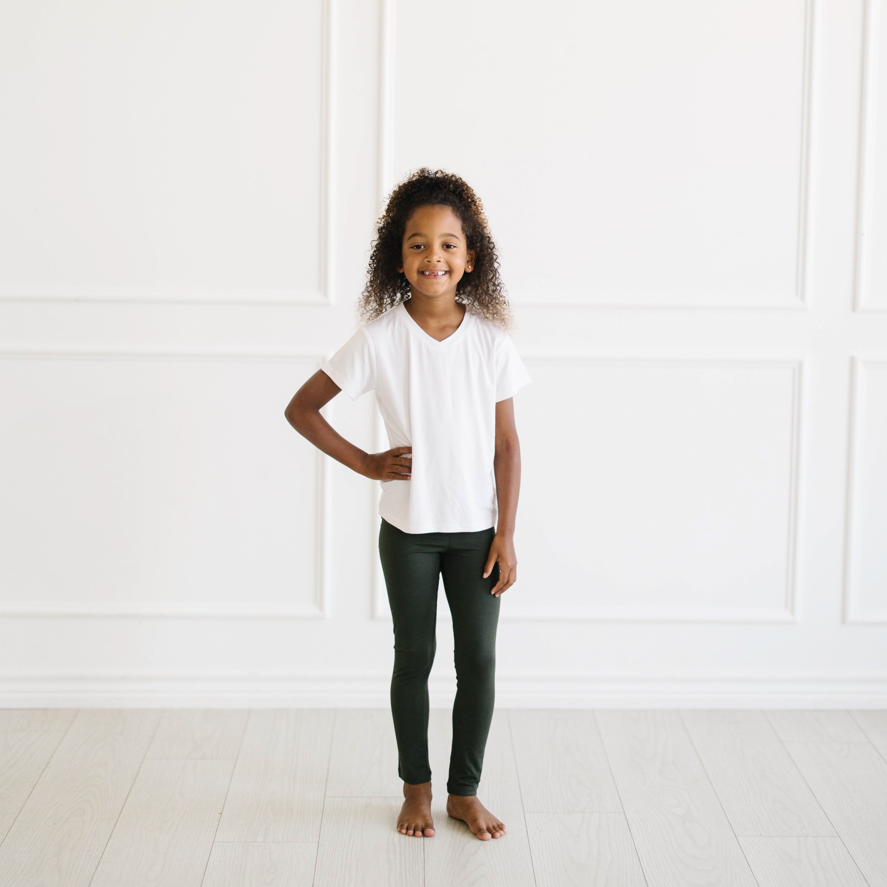 Young girl wearing Fir Legging and Snow V-neck tee standing in a minimalistic room with white walls and floor.