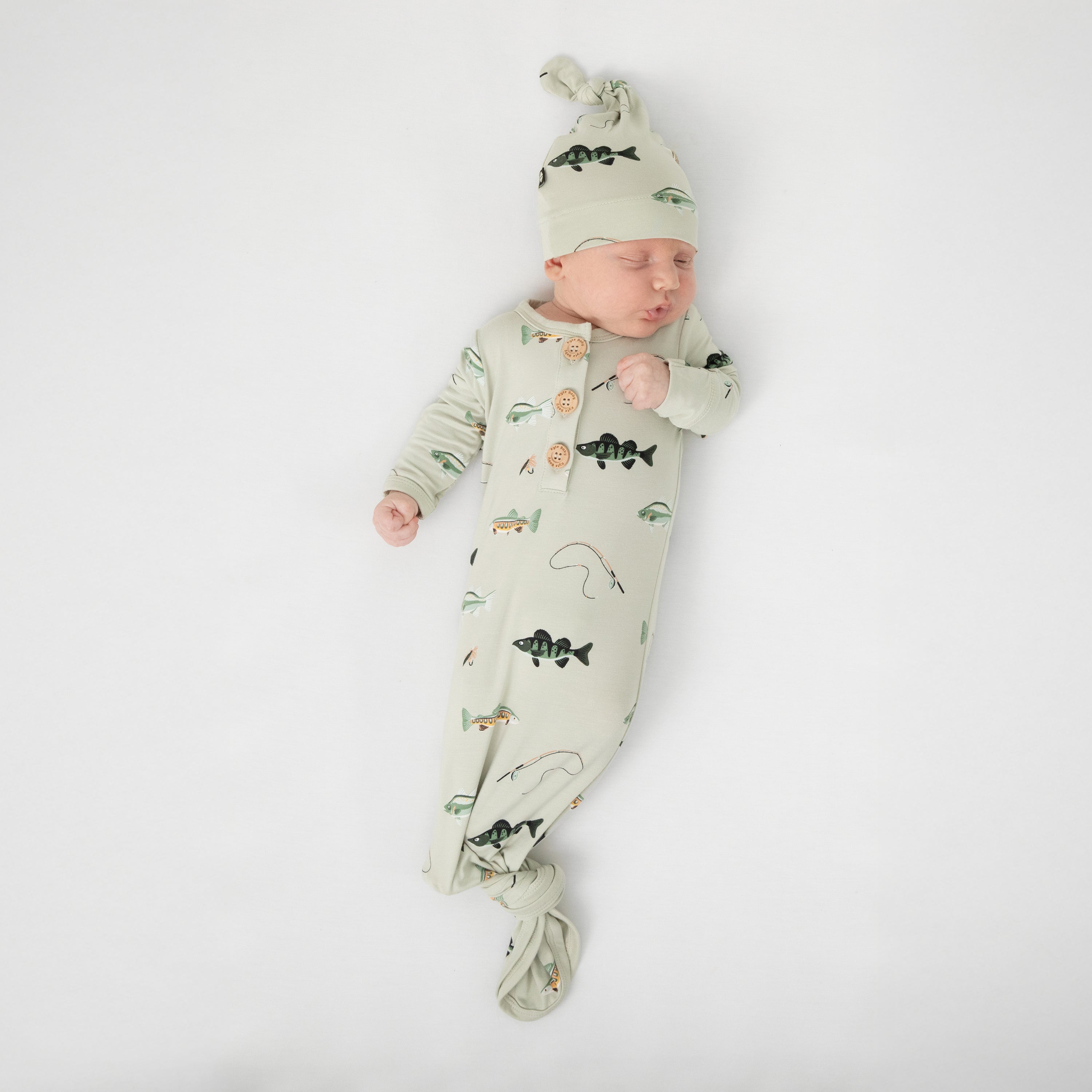 Sleeping newborn laying on a white blanket wearing the Knotted Gown with Hat Set in Fishing tied at the bottom