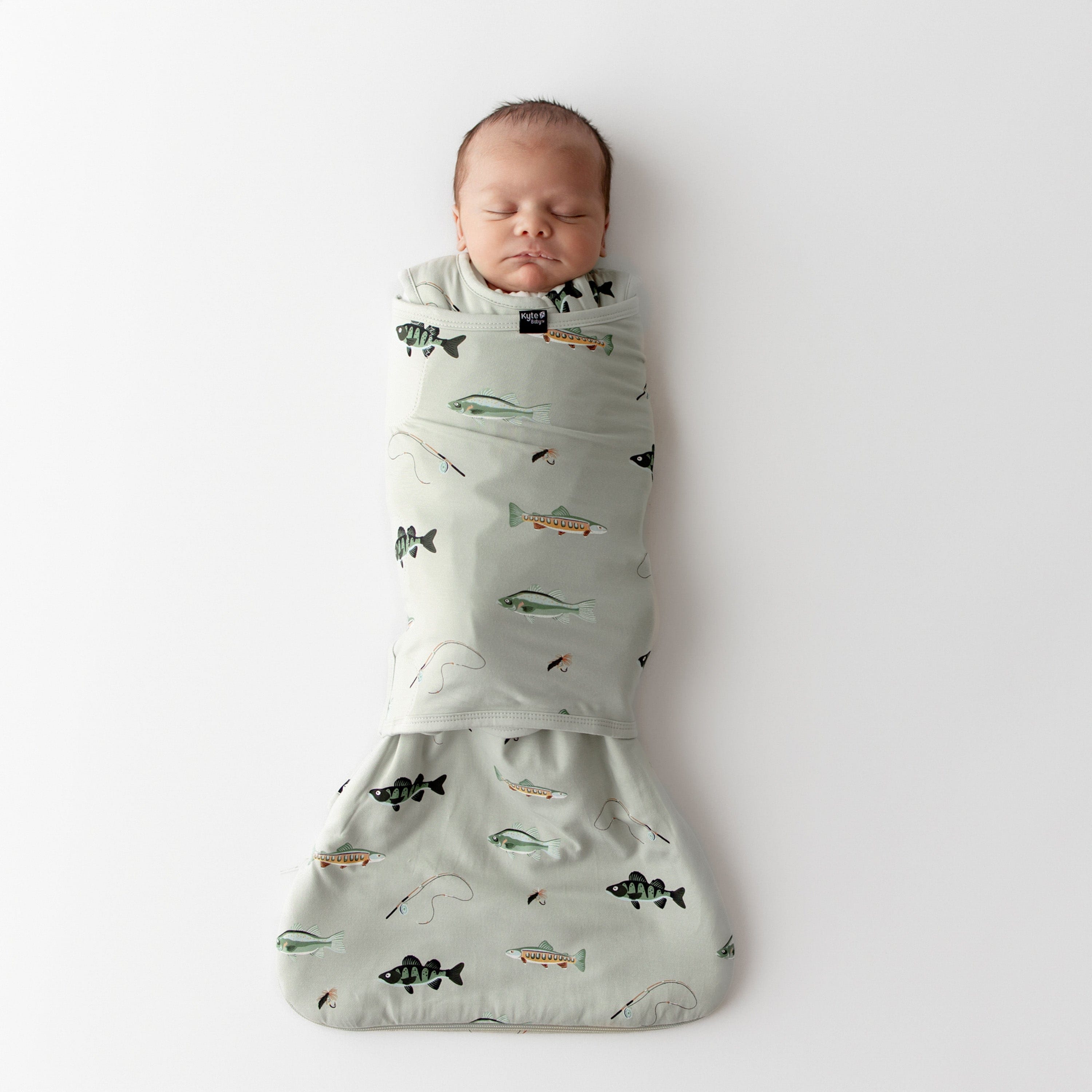 Newborn baby swaddled in a green sleep bag swaddler with fishing prints on a white background