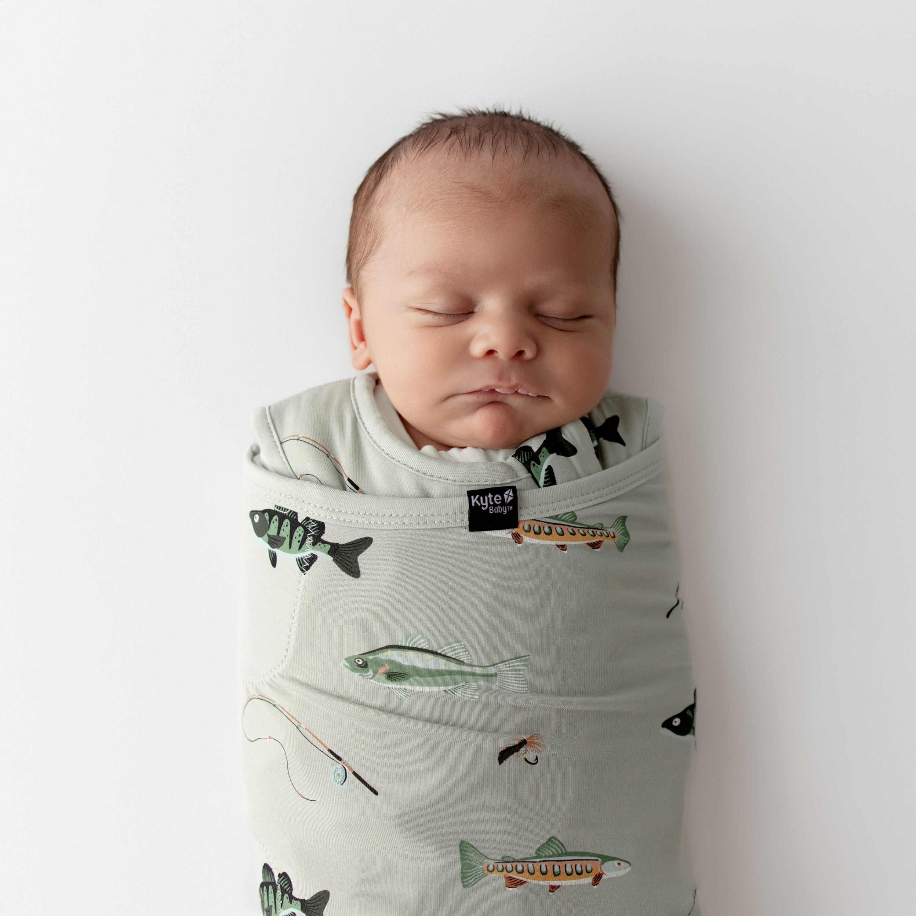 Newborn baby swaddled in a green sleep bag swaddler with fishing prints on a white background