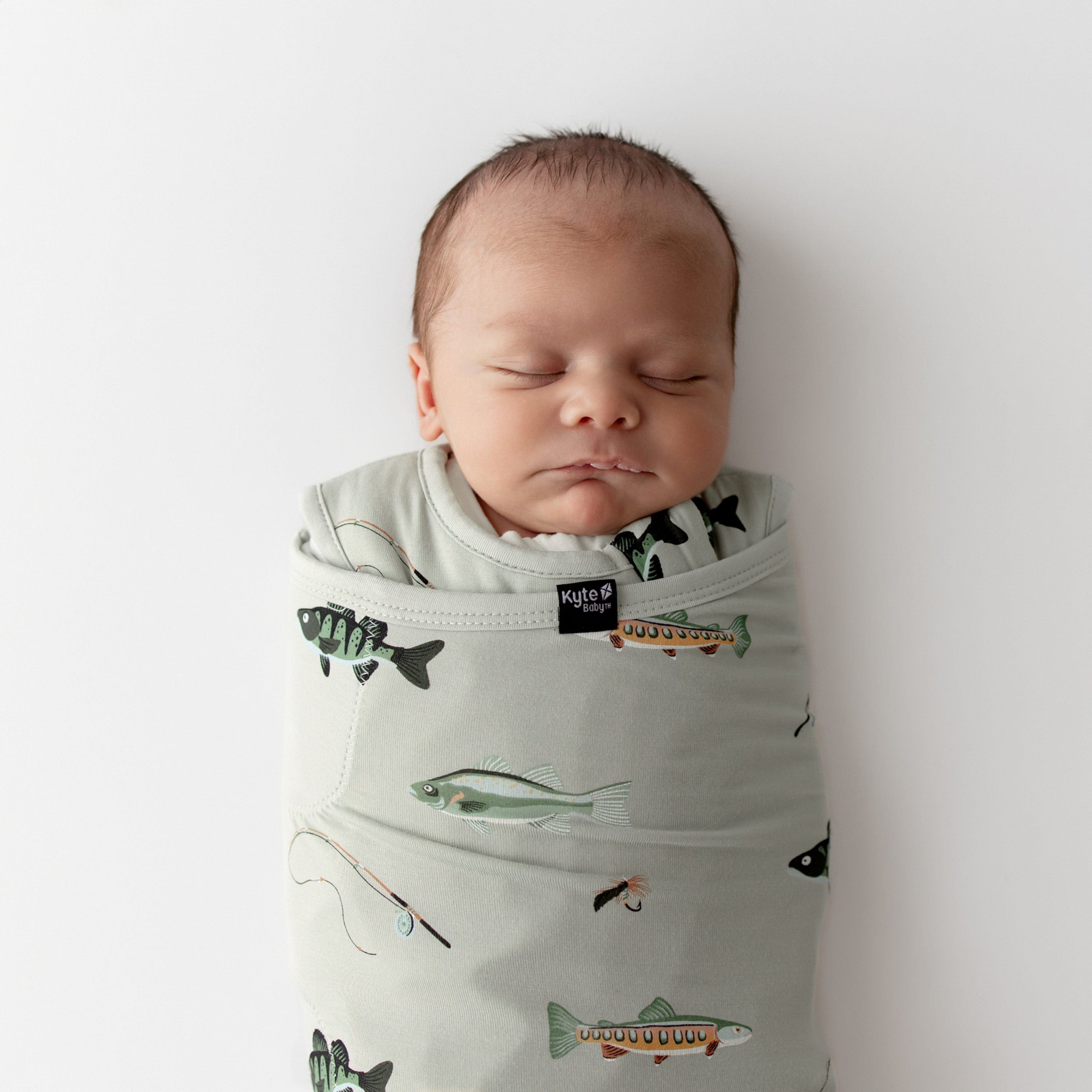 Newborn baby swaddled in a green sleep bag swaddler with fishing prints on a white background
