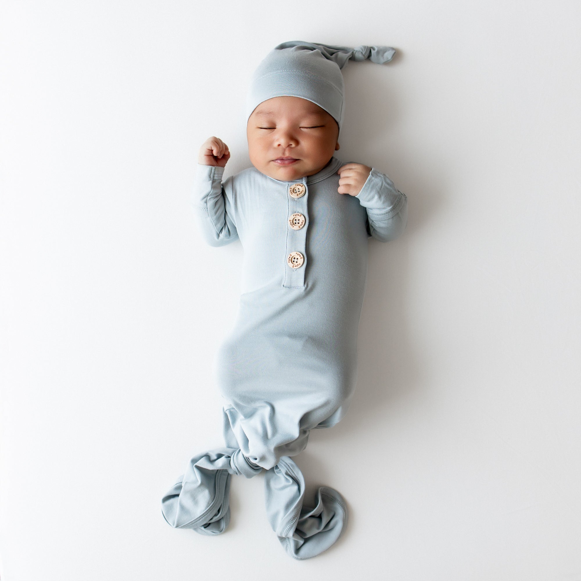Baby asleep wearing Kyte Baby knotted gown and hat set in a dusty blue color on a white backdrop