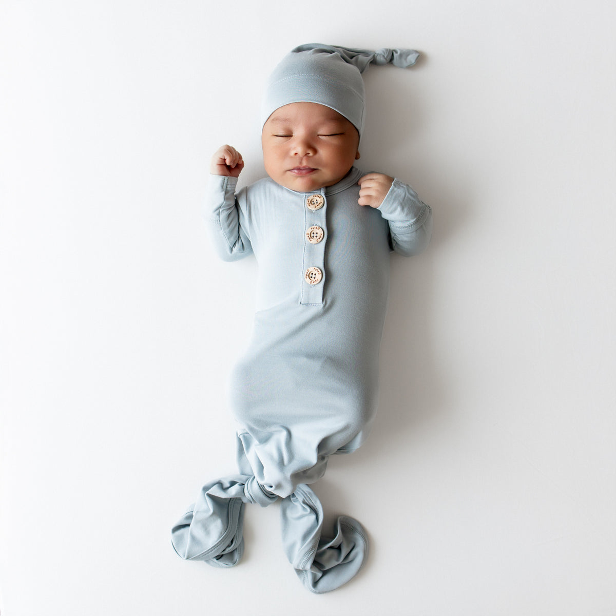 Baby asleep wearing Kyte Baby knotted gown and hat set in a dusty blue color on a white backdrop