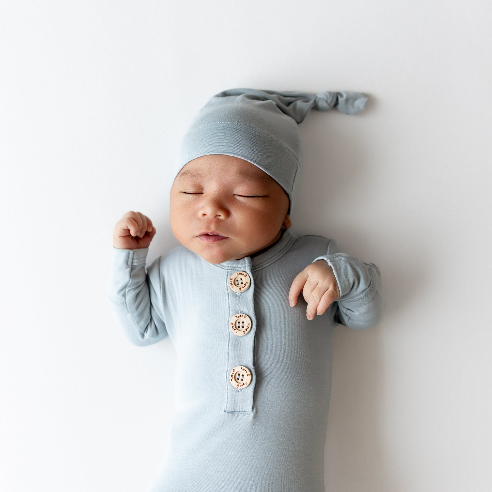 Baby asleep wearing Kyte Baby knotted gown and hat set in a dusty blue color with wooden buttons on a white backdrop