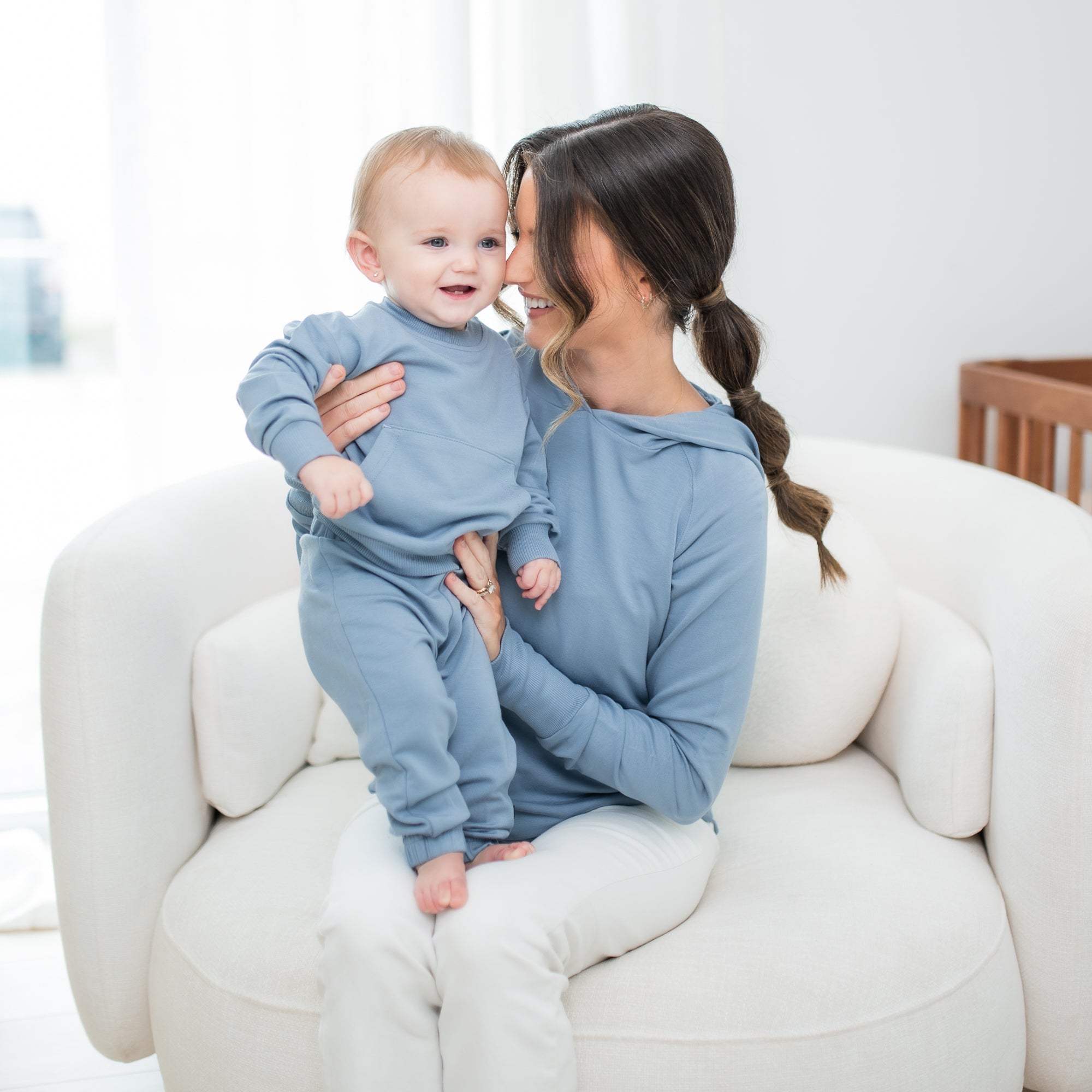 Mother and child sitting other on a cream colored sofa chair matching in Slate. Young toddler wearing the French Terry Jogger Set in Slate with mom wearing the French Terry nursing Hoodie