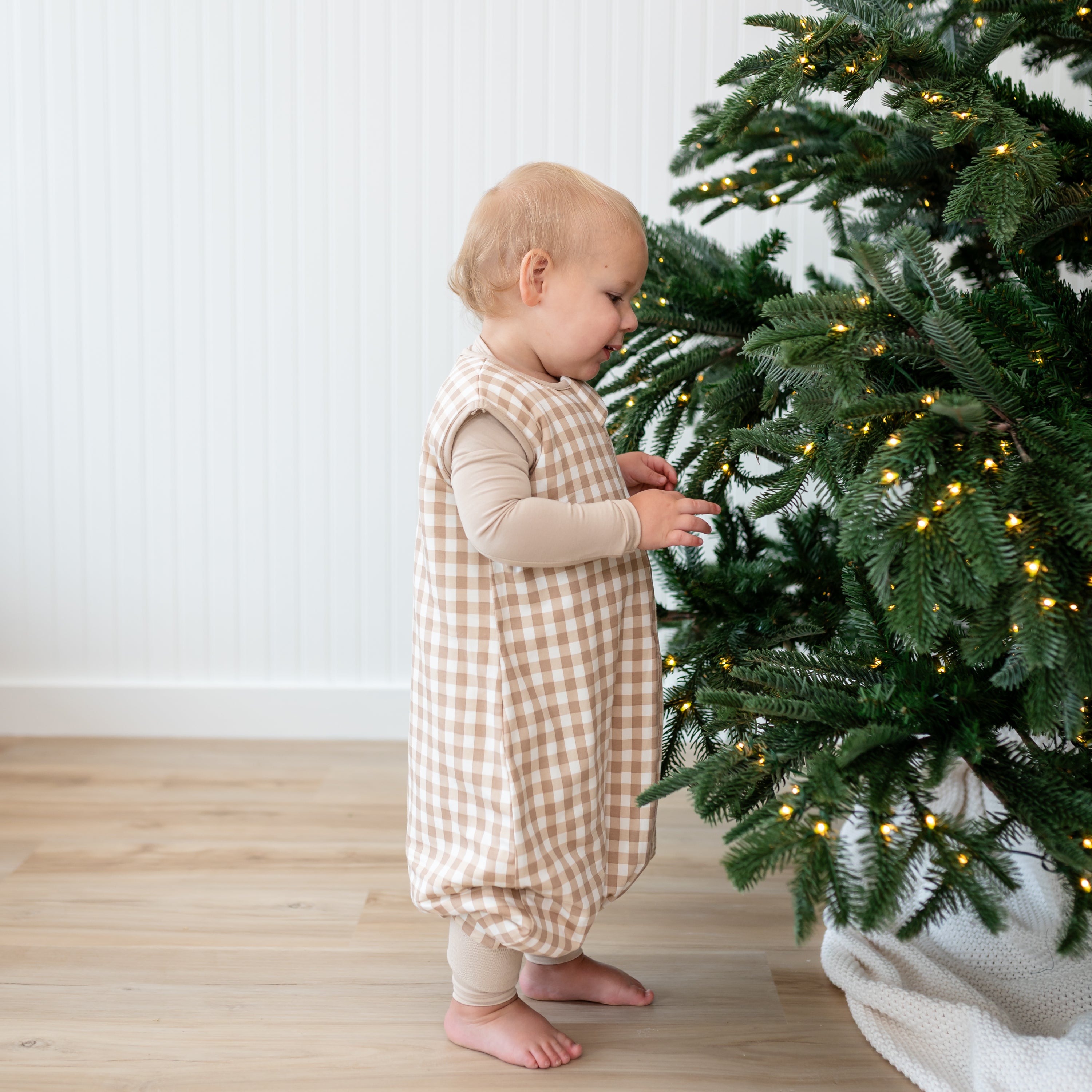 Toddler standing beside a Christmas Tree wearing the Sleep Bag Walker in Gingham Bisque 1.0