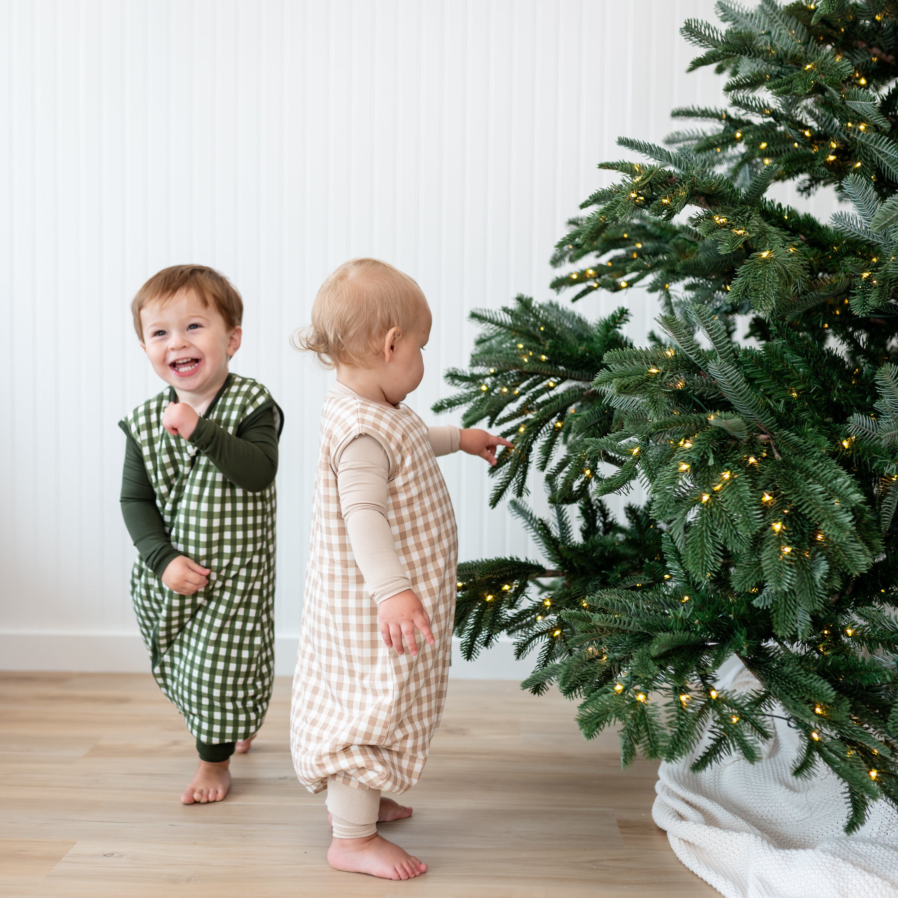 Two toddlers wearing the Sleep Bag Walker in Gingham Bisque 1.0 and Gingham Fir 1.0 beside a Christmas Tree