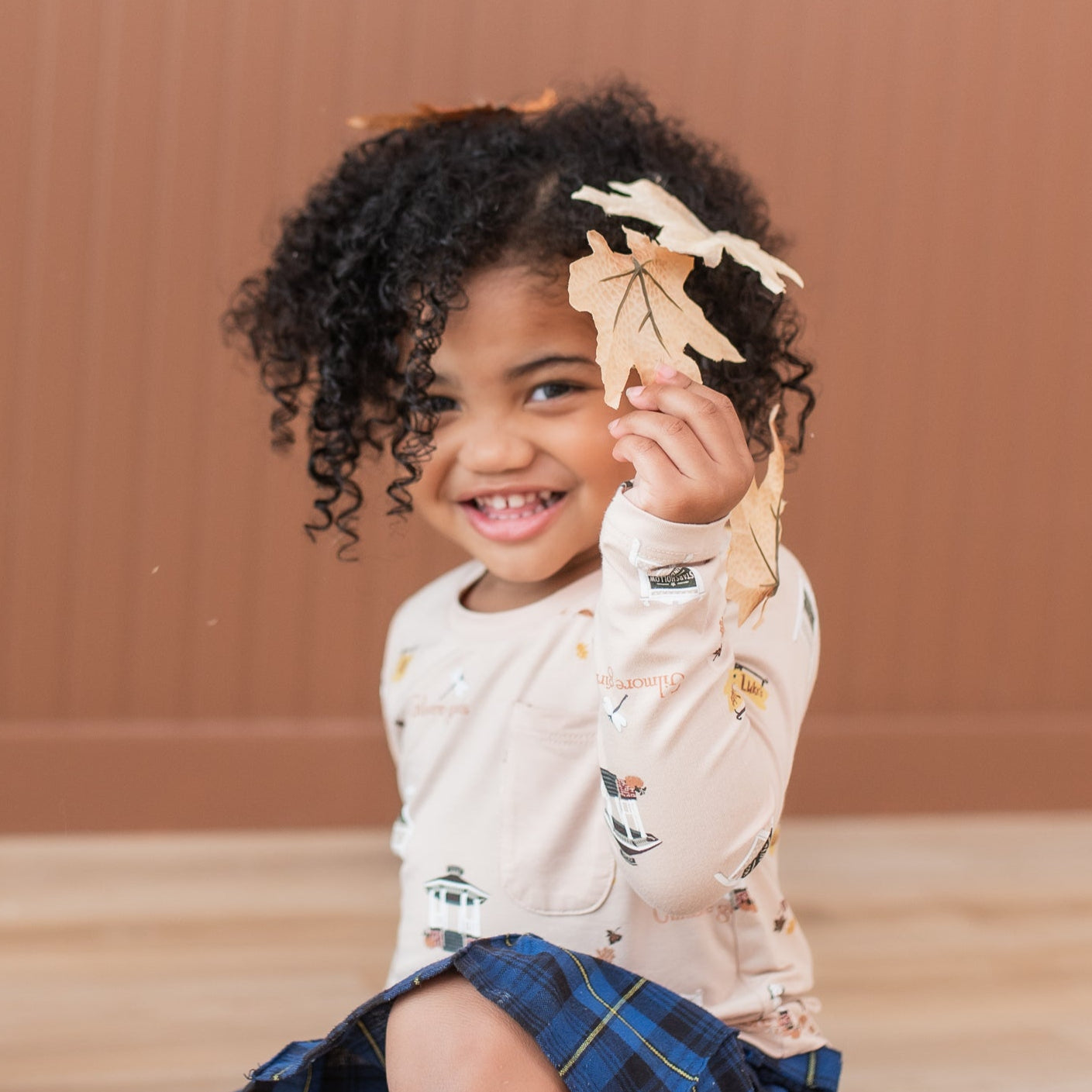 Young girl sitting on the floor holding up a leaves wearing the Long Sleeve Toddler Crew Neck Tee in Gilmore Girls