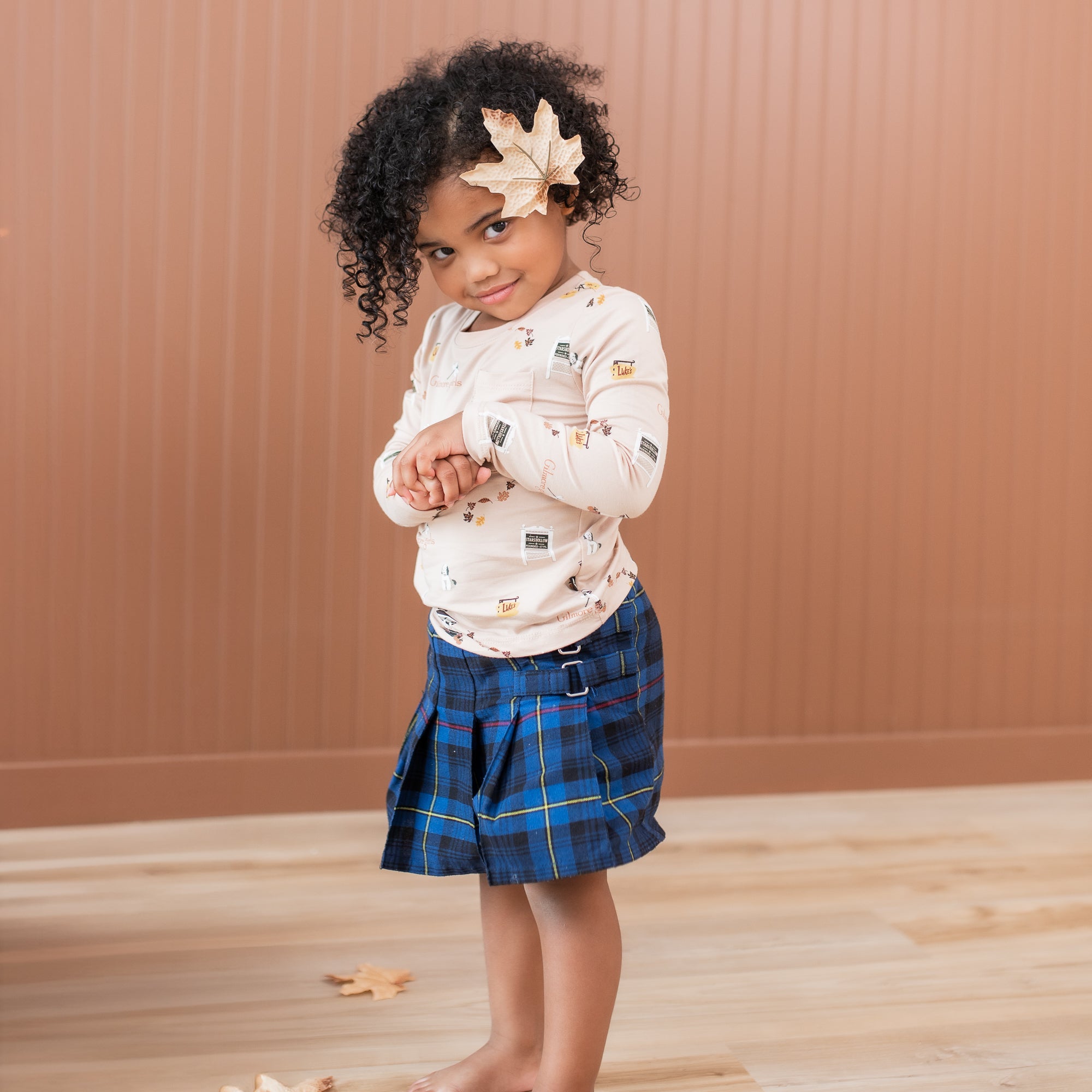 Young girl standing wearing the Long Sleeve Toddler Crew Neck Tee in Gilmore Girls with a blue plaid skirt with a leaf in her hair