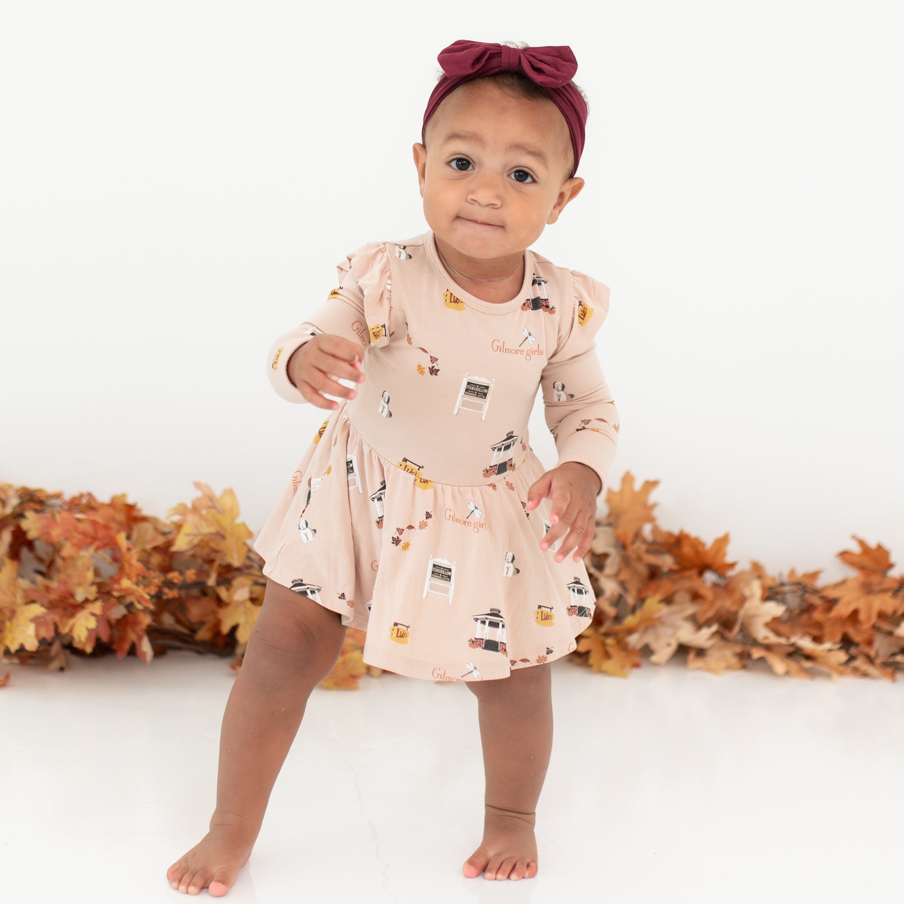 Young toddler walking wearing the Long Sleeve Twirl Bodysuit Dress in Gilmore Girls with a burgundy bow in front of a leaf garland and white backdrop