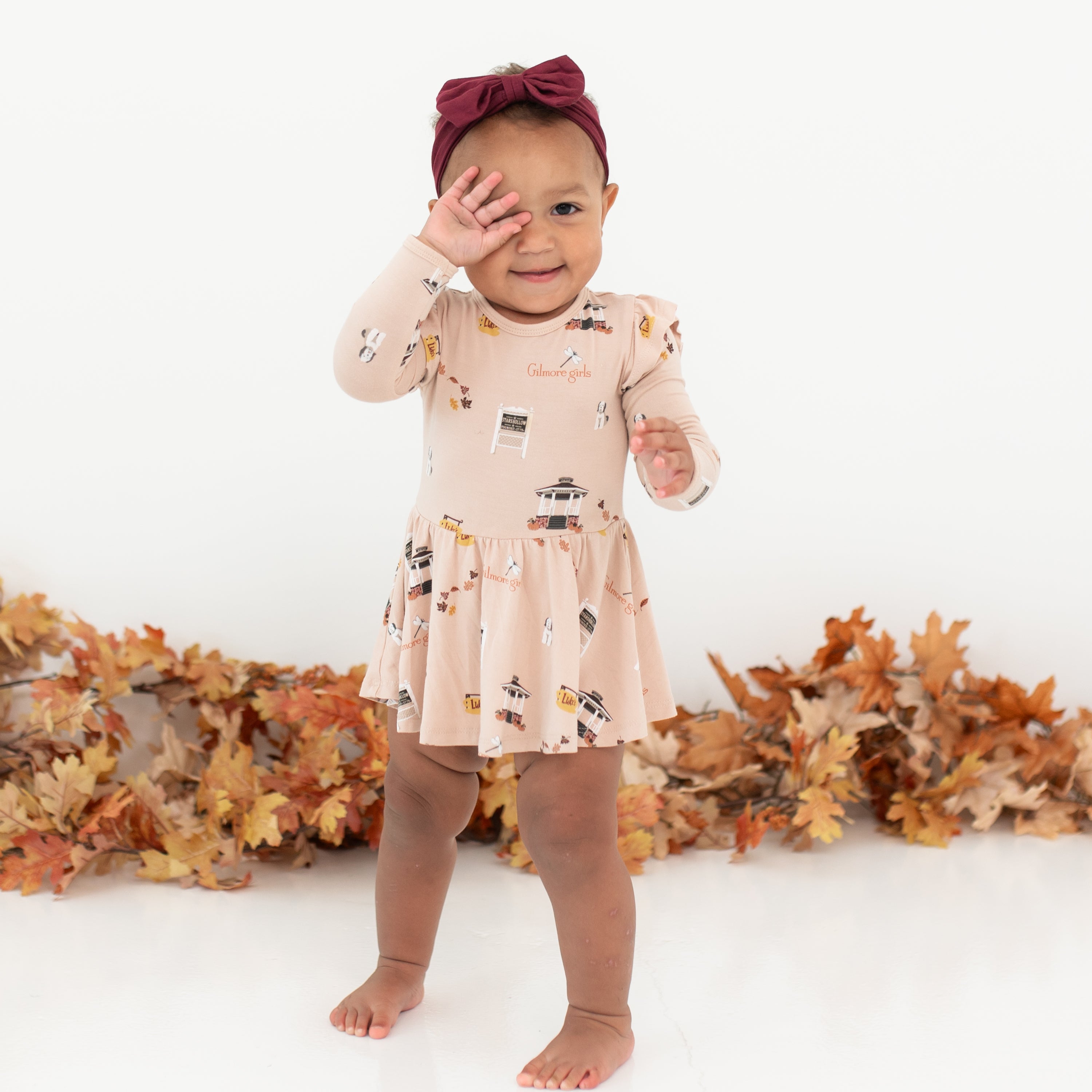 Young toddler covering her eye with one hand wearing the Long Sleeve Twirl Bodysuit Dress in Gilmore Girls and a burgundy bow standing in front of a white backdrop and leaf garland