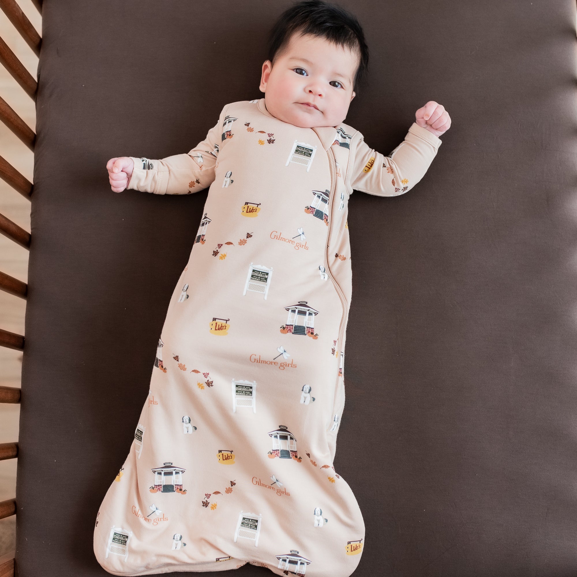 Infant laying down in a crib on an Espresso crib sheet wearing the Sleep Bag in Gilmore Girls 1.0 with a matching romper