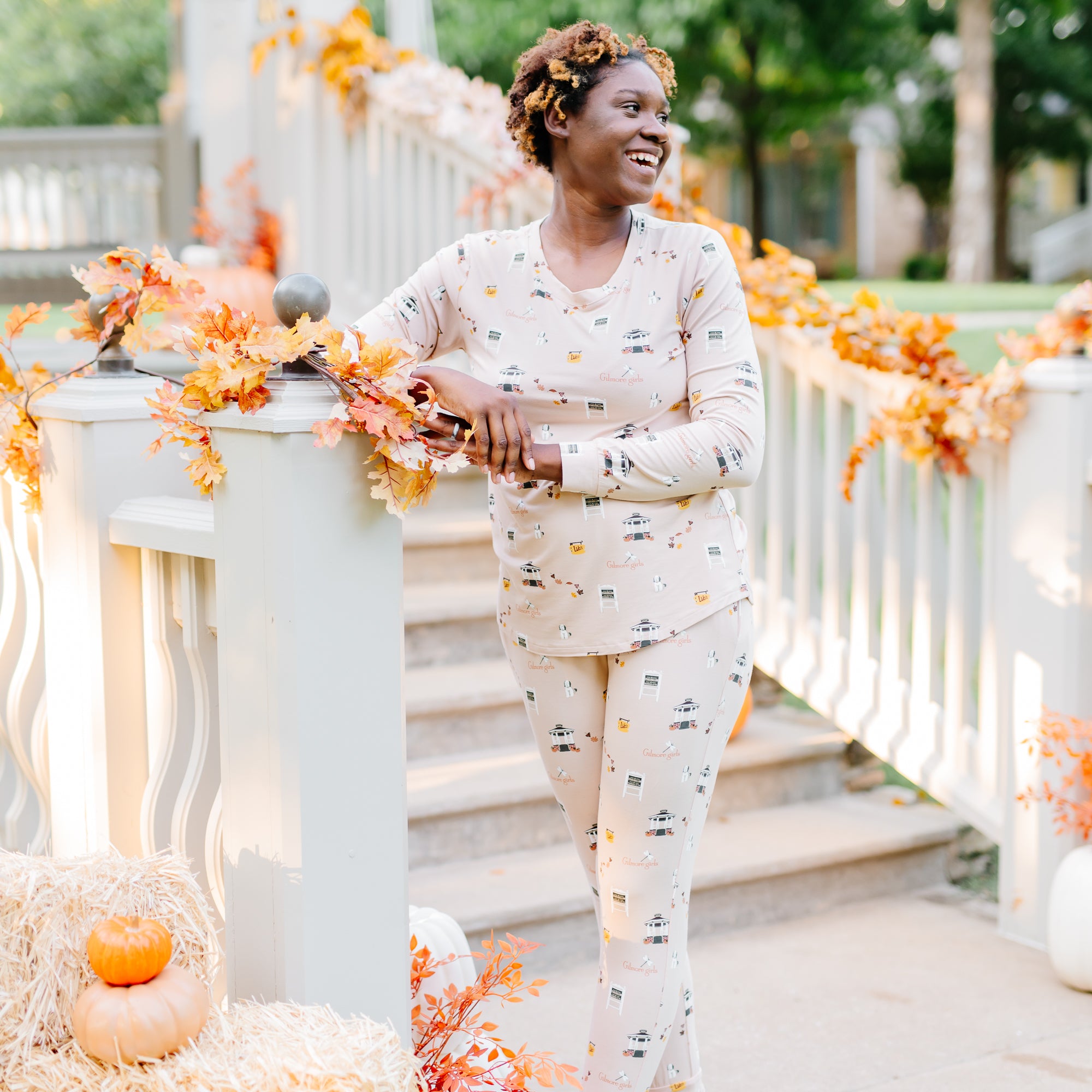 Smiling model leaning against the white railing leading up to a decorated fall gazebo wearing the Women's Jogger Pajama Set in Gilmore Girls
