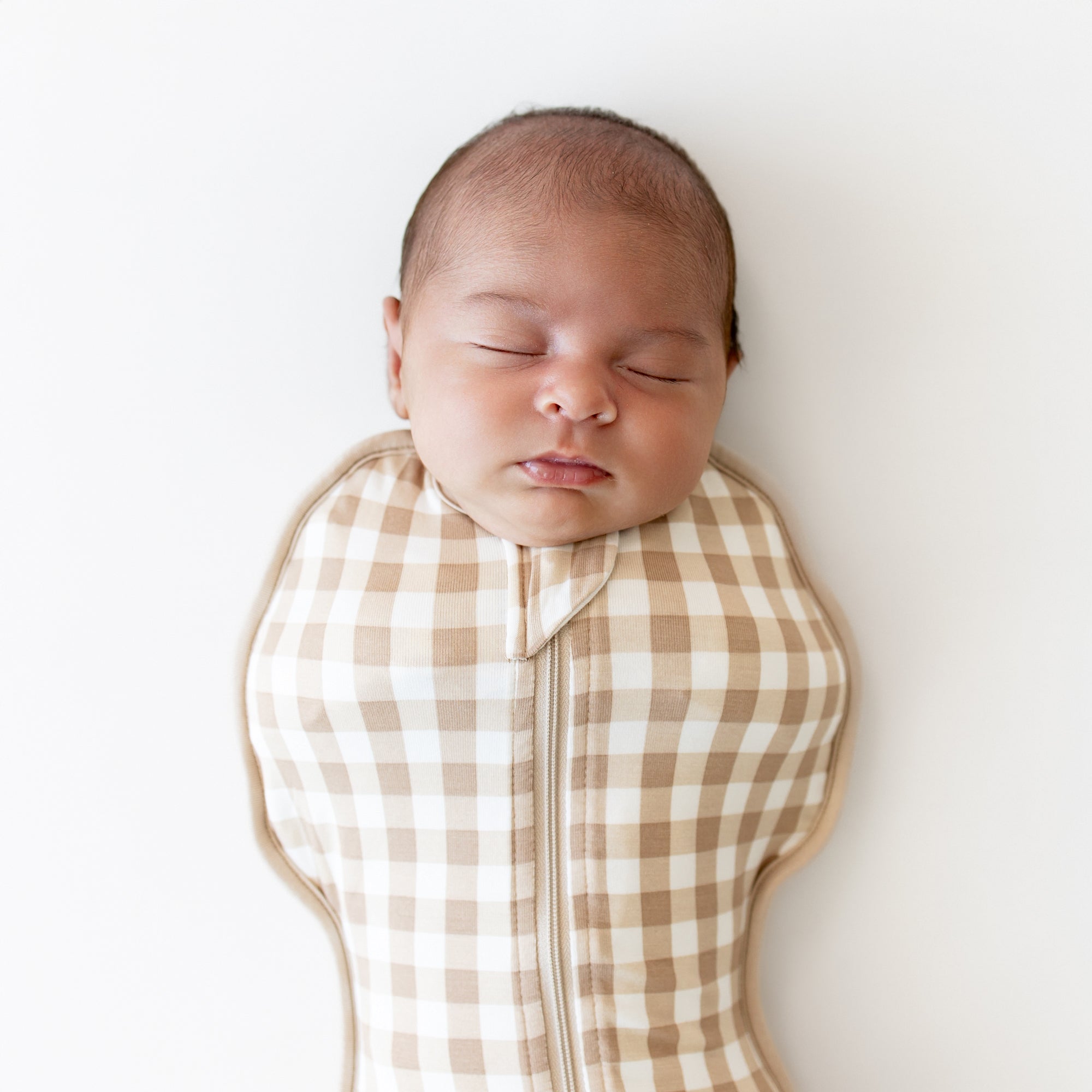 Newborn baby swaddled in a brown and white checkered swaddle bag on a white background